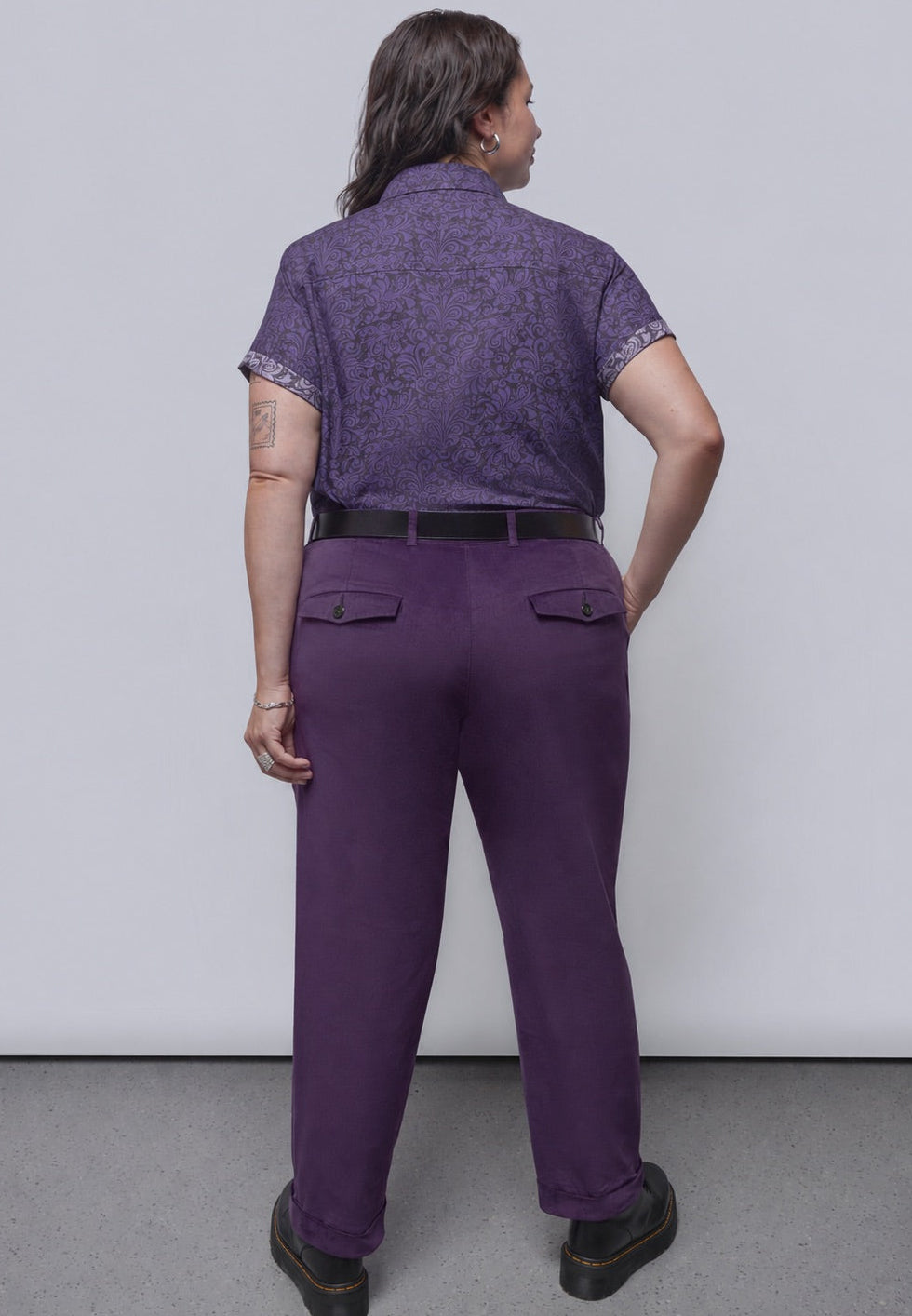 1X=A person shown from the back wearing a purple short-sleeve patterned shirt and matching purple trousers, standing against a plain light gray wall and gray floor.