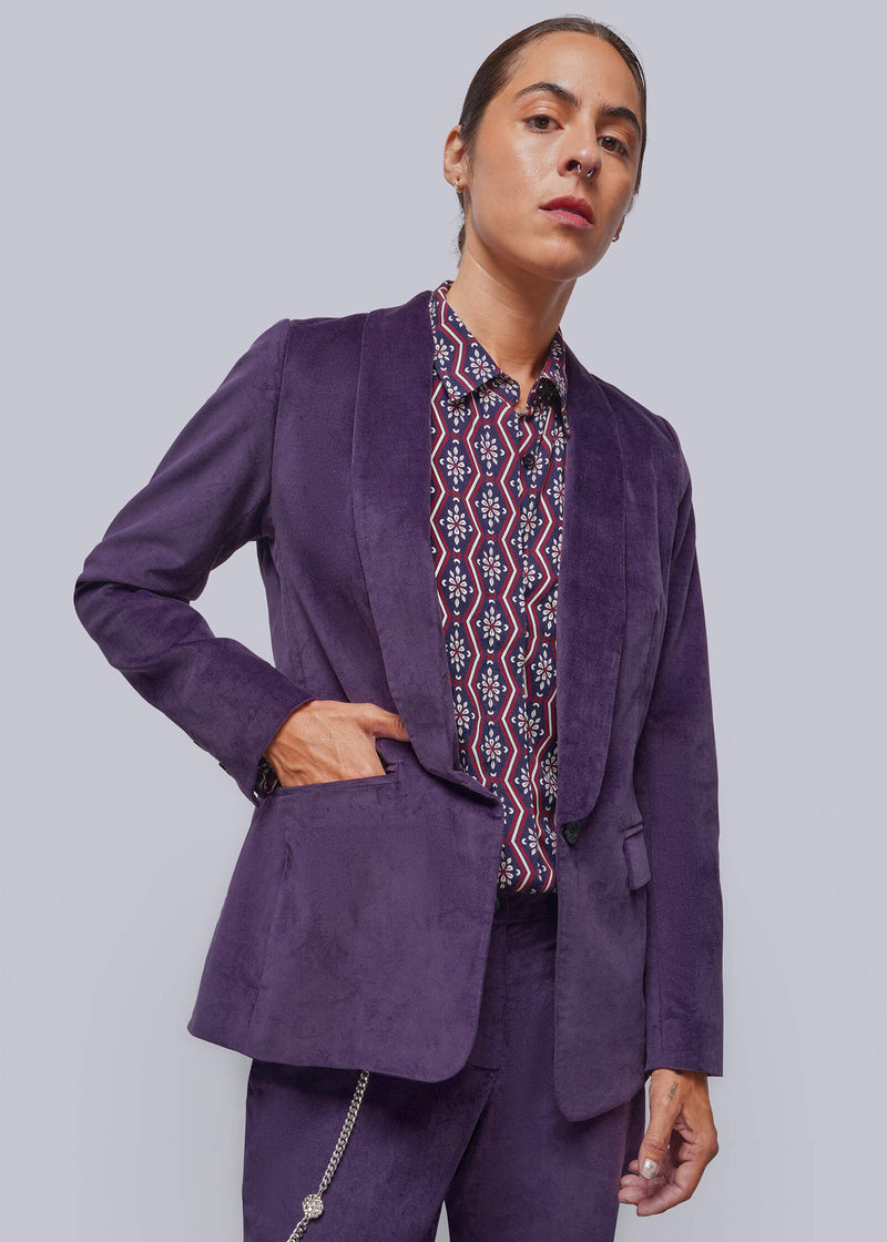 S=A model wearing a purple velvet blazer and matching trousers, styled with a patterned button-down shirt, against a solid gray studio background.