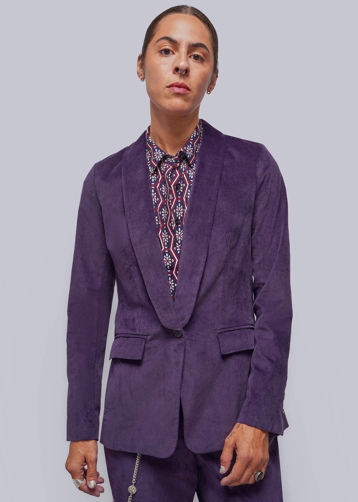 S=A model wearing a purple corduroy blazer and matching trousers, styled with a patterned shirt, against a solid gray background.