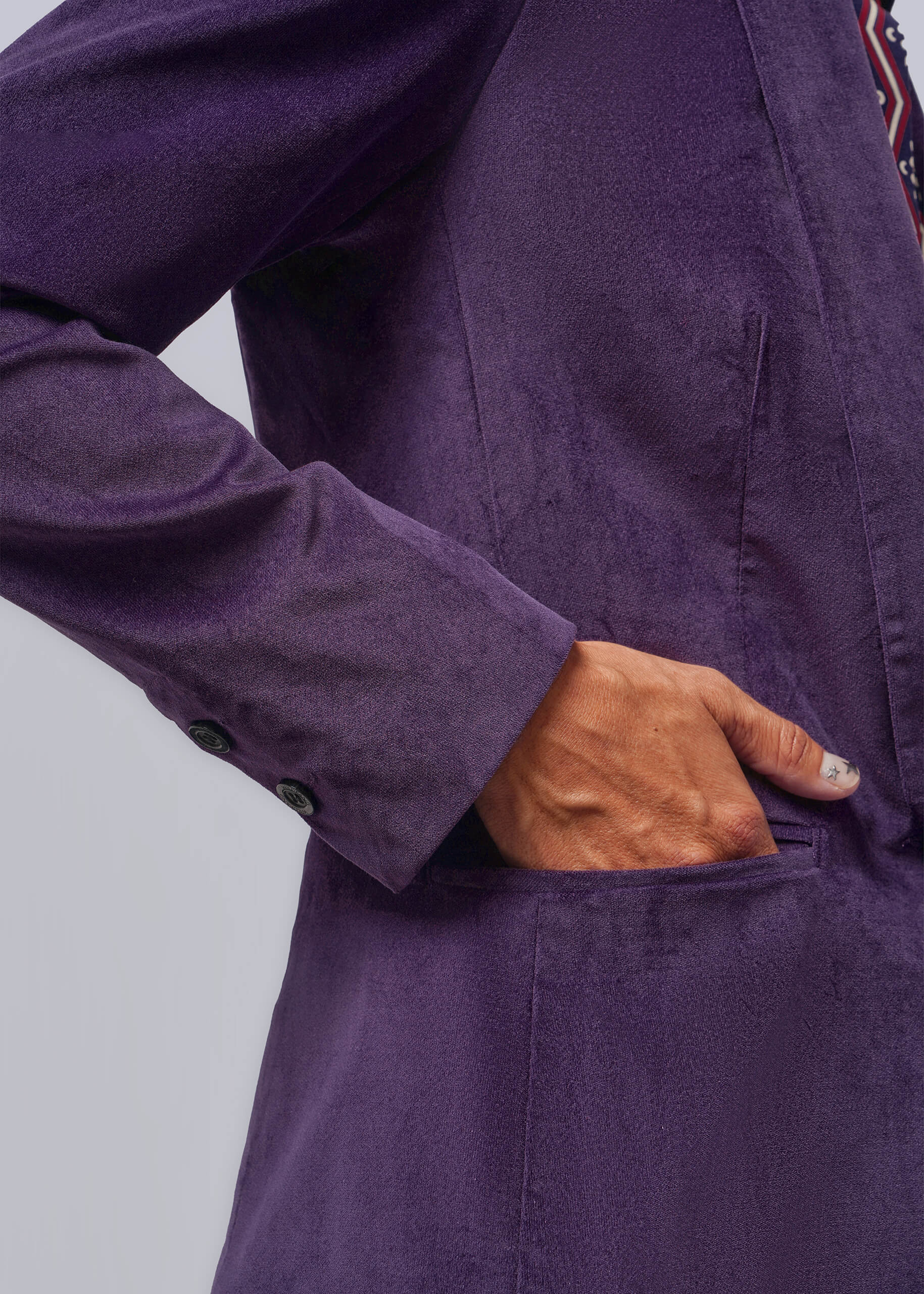 Close-up of a person wearing a textured purple blazer, with one hand tucked into a front pocket. The fabric has a velvet texture, and a patterned shirt is partially visible at the collar against a light gray background.