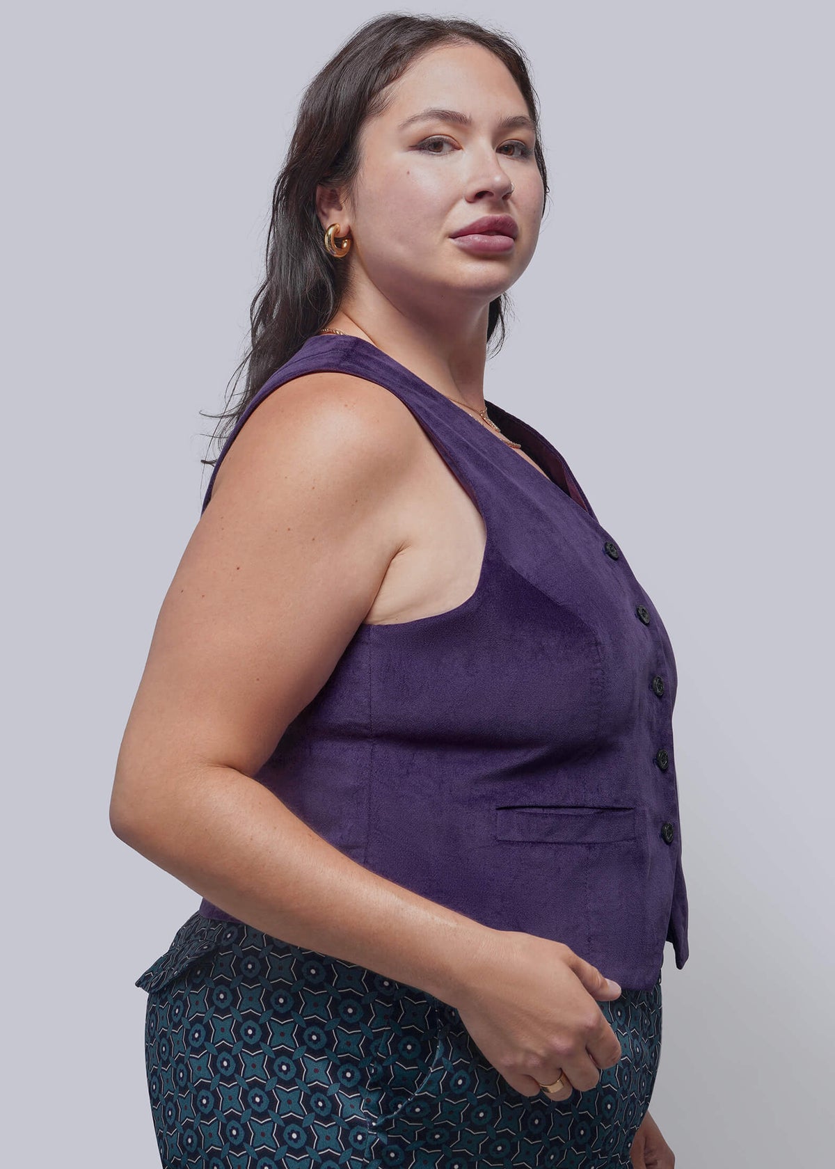 1X=A woman wearing a sleeveless purple vest with dark buttons and a matching purple top, paired with a patterned skirt, photographed against a solid gray studio background.