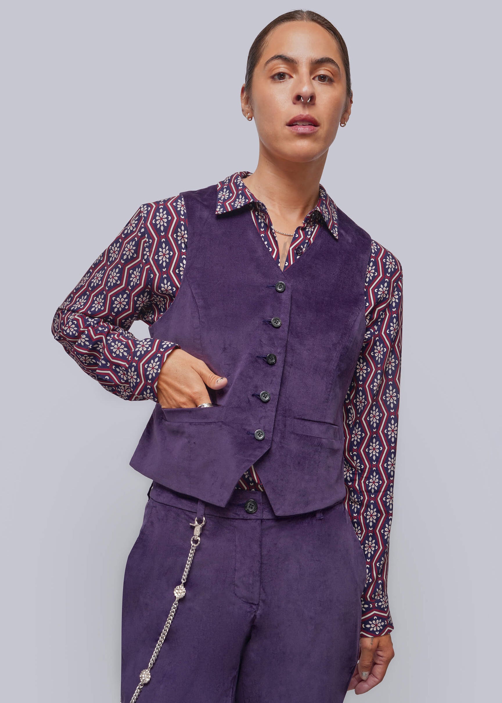 A model wearing a purple velvet vest and matching trousers over a patterned long-sleeve shirt, posed against a solid gray studio background.