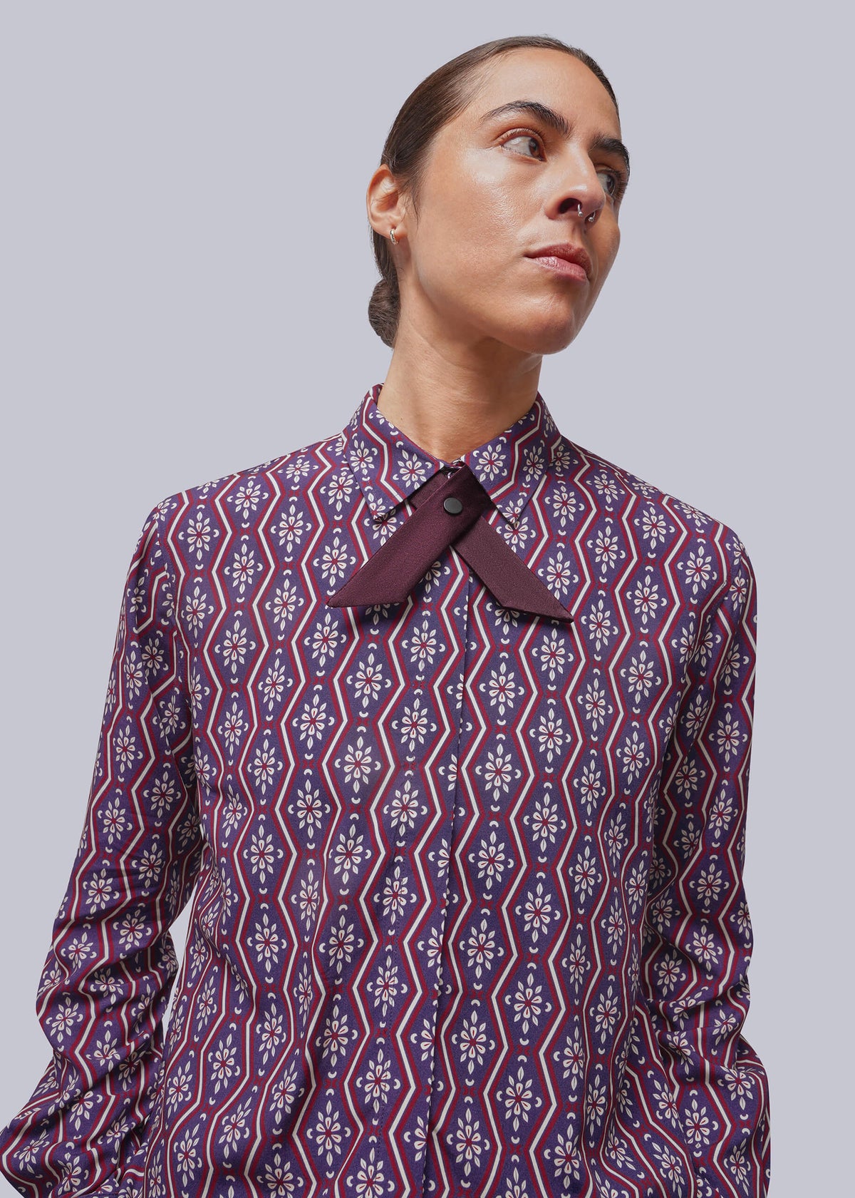 A person wearing a long-sleeve button-up shirt with a dark purple and navy blue geometric pattern and a contrasting dark purple bow tie at the collar, set against a solid gray background.