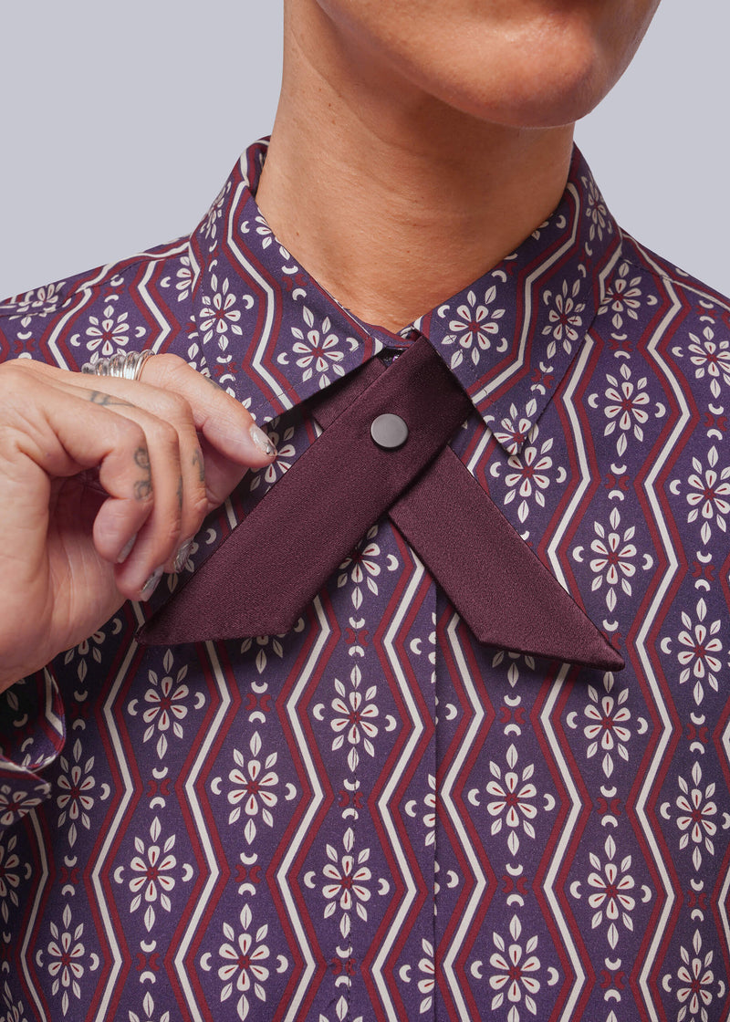 Close-up of a person wearing a patterned button-up shirt with a contrasting dark burgundy bow tie and a hand adjusting it. The shirt features a purple, red, and cream geometric floral pattern.