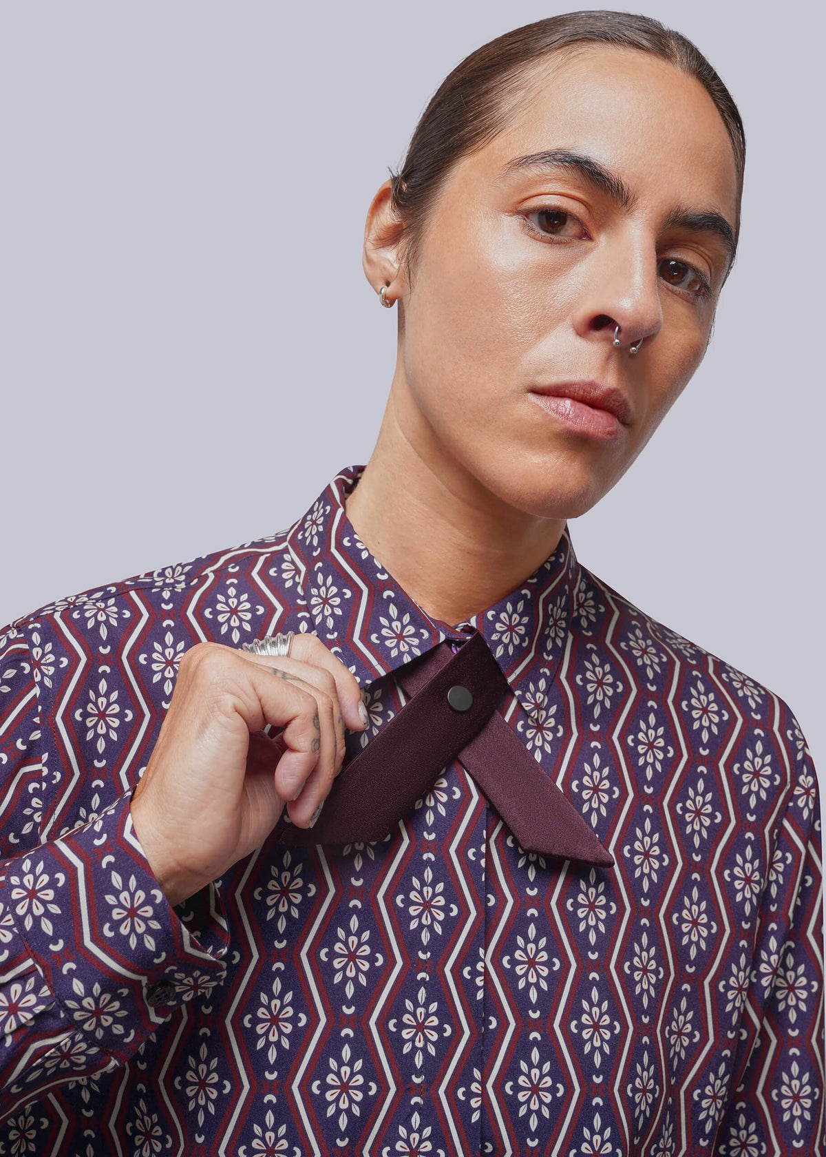 A close-up of a model wearing a patterned button-down shirt with a bow tie around the collar. The shirt features a geometric and floral pattern in navy blue, burgundy, and cream.