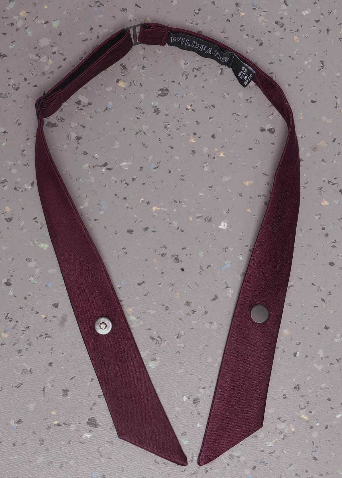 A burgundy satin necktie with a wide collar, laid flat on a speckled gray concrete surface. The tie features silver-tone metal snap buttons on the front and a black fabric neck strap with a silver metal buckle.