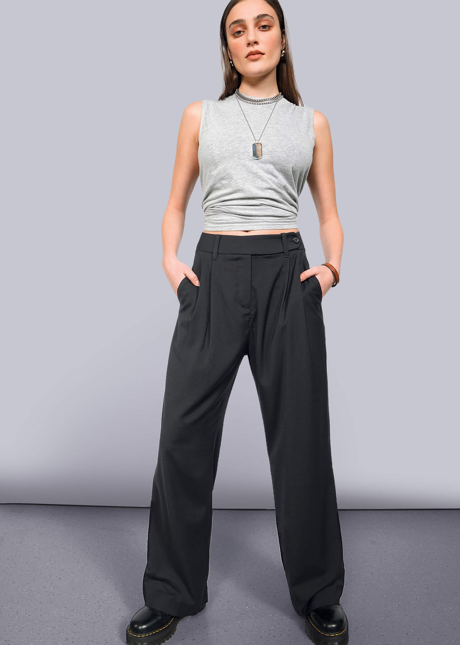 S=A person stands confidently against a white background, wearing The Empower Wide Leg Trouser with high waist and pleated front, paired with a gray sleeveless crop top, black shoes, layered necklaces, and hands in their pockets.