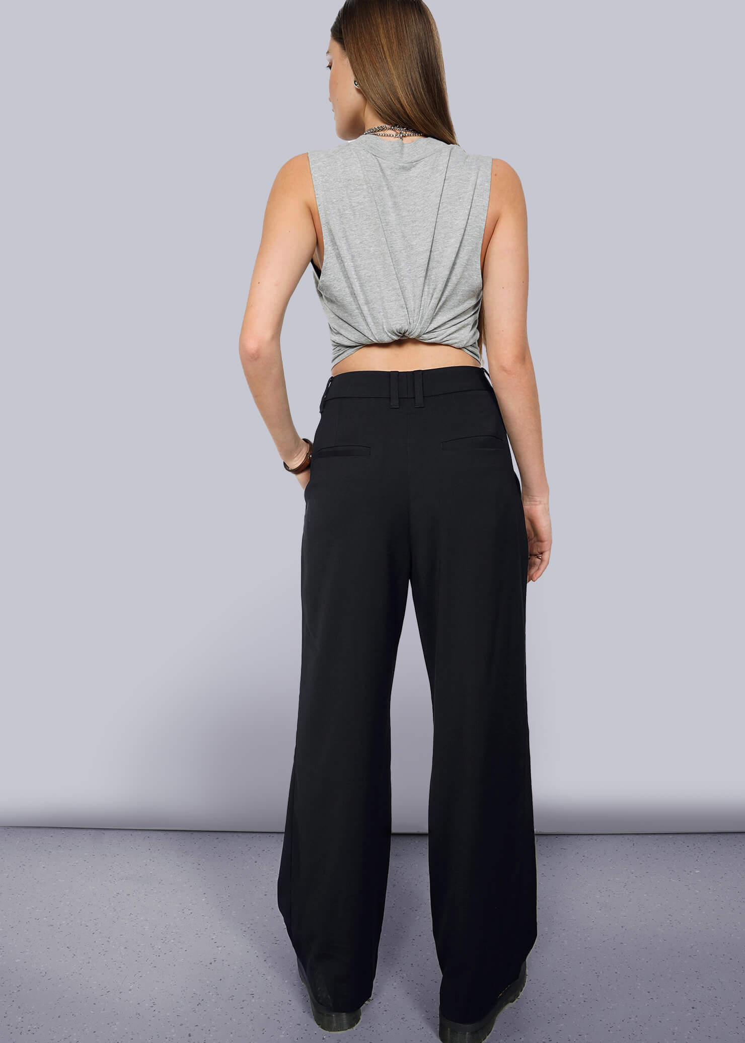 S=A person with long brown hair stands with their back to the camera, wearing a sleeveless gray top in a knot and The Empower Wide Leg Trouser for a stylish, modern look.
