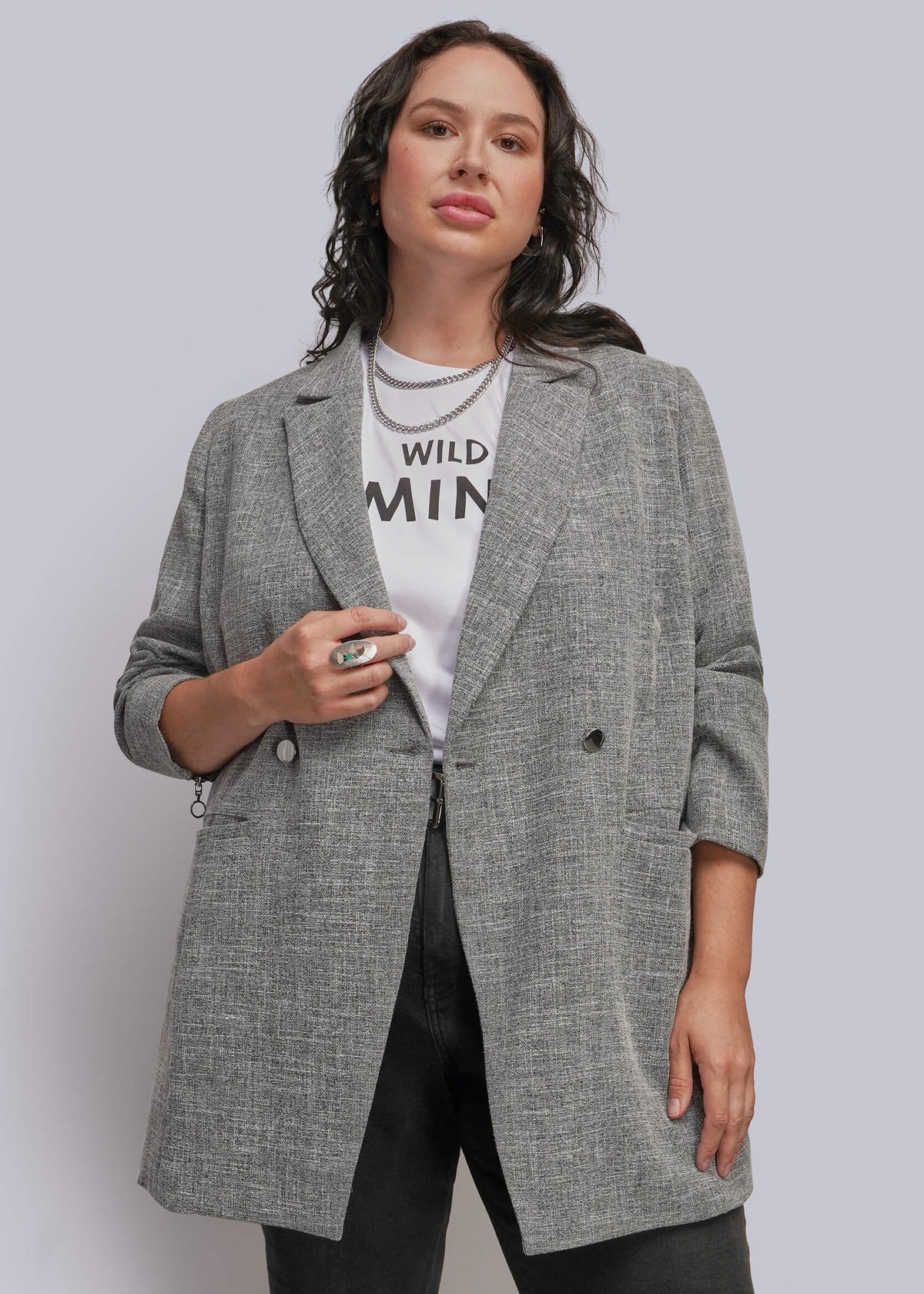 1X=A woman modeling a double-breasted gray textured blazer over a white graphic t-shirt and black pants, standing against a light gray studio background.