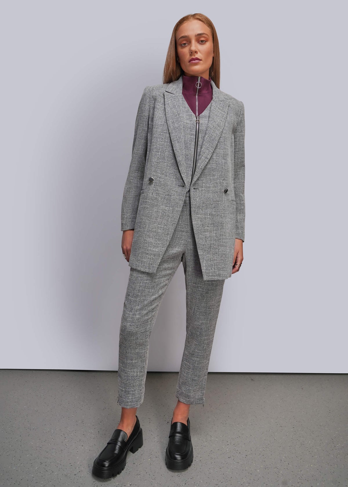 S=A woman modeling a tailored gray suit with a blazer and cropped trousers, worn over a dark purple mock neck top and black loafers, standing against a light gray studio wall.