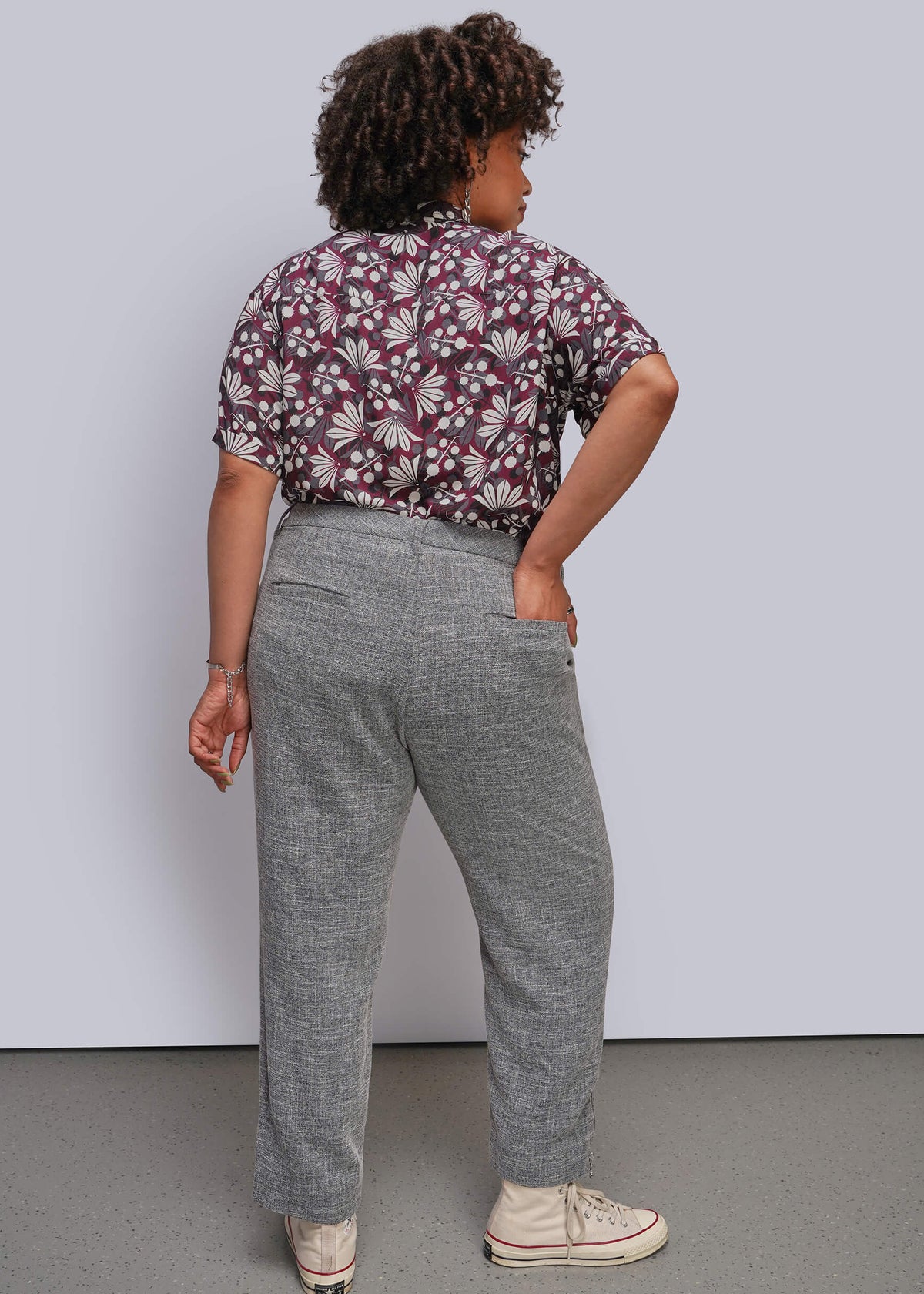 1X=A model is shown from the back wearing a patterned short-sleeve button-up shirt in burgundy and white, paired with gray trousers and beige sneakers, set against a plain light gray studio background.