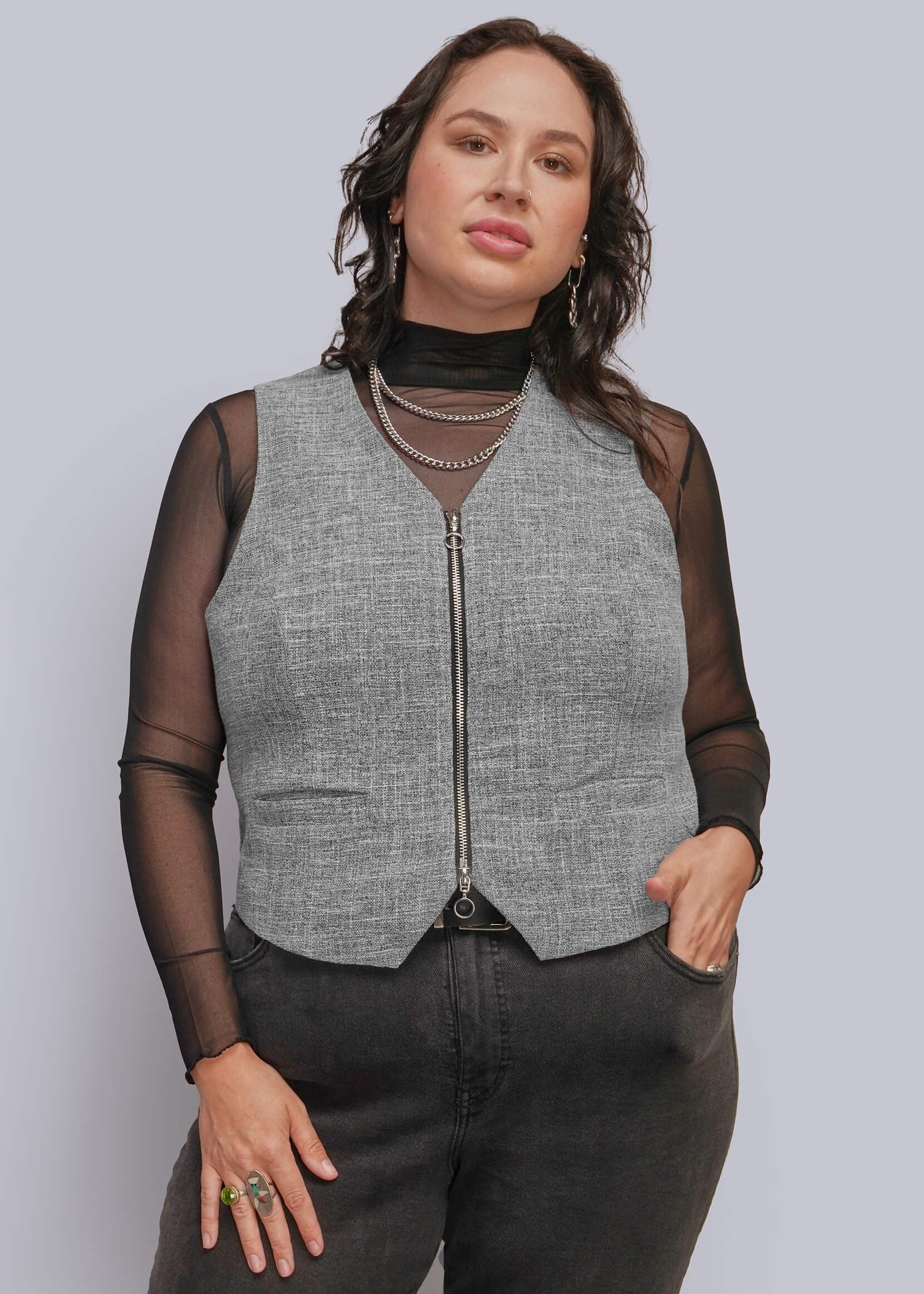 1X=A person modeling a sleeveless gray textured zip-front vest over sheer black long-sleeve mesh top, paired with dark gray jeans and layered necklaces, against a light gray studio background.