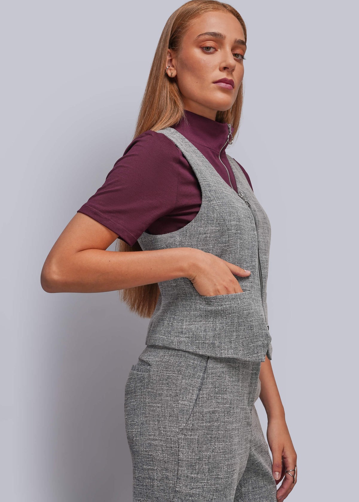 S=A woman modeling a gray two-piece vest and trouser set over a deep purple mock-neck t-shirt, photographed against a solid light gray studio background.