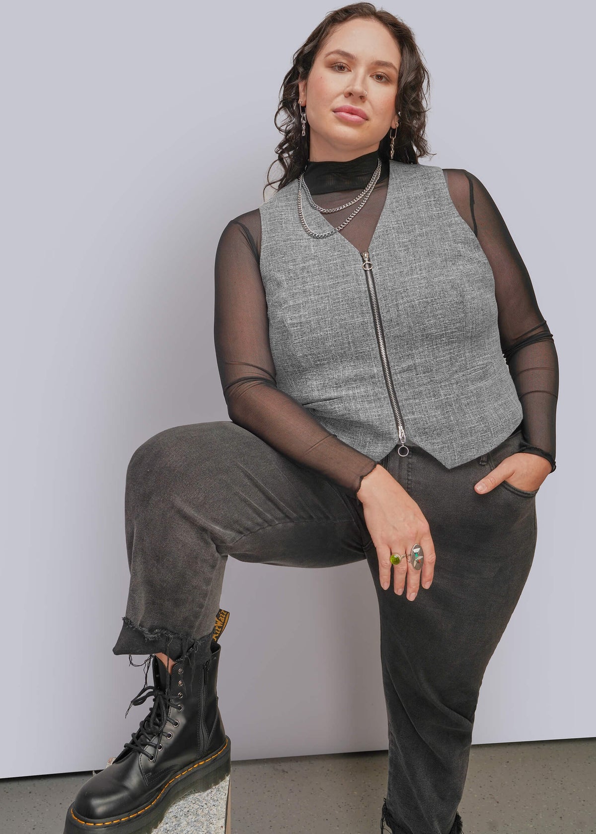 1X=A person models a sleeveless gray zip-front vest over sheer black long-sleeve mesh top, paired with dark charcoal pants and black lace-up boots, seated against a light gray studio background.