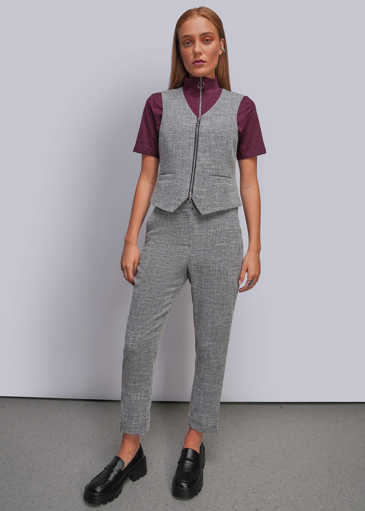 S=A model stands against a light gray studio wall wearing a coordinated gray vest and trouser set over a burgundy short-sleeve mock-neck top, paired with black loafers.