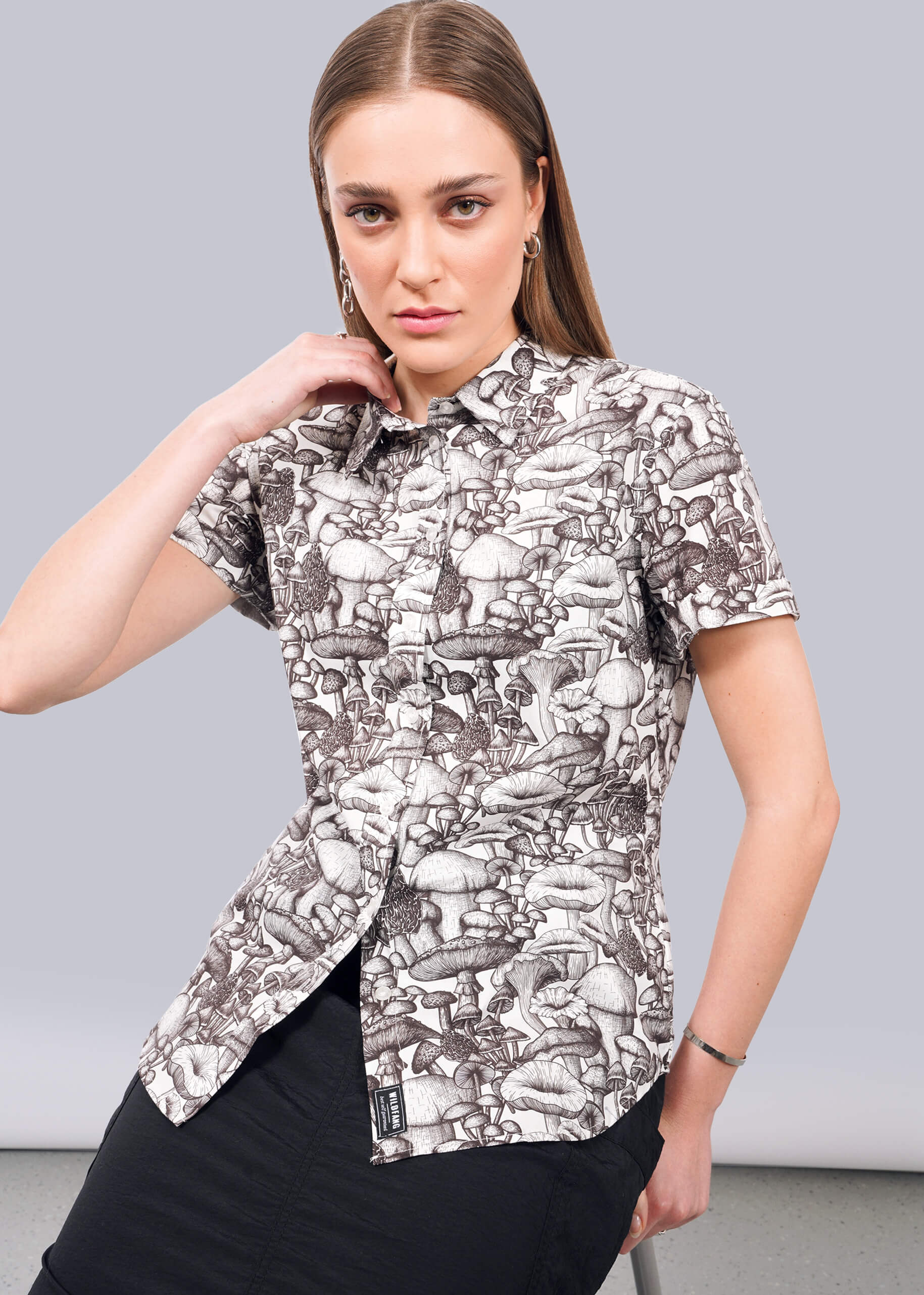 S= A woman with straight brown hair wears The Essential Button Up, a tailored-fit, short-sleeved shirt in breathable cotton featuring a black and white mushroom pattern. She sits against a plain, light background with a serious expression.