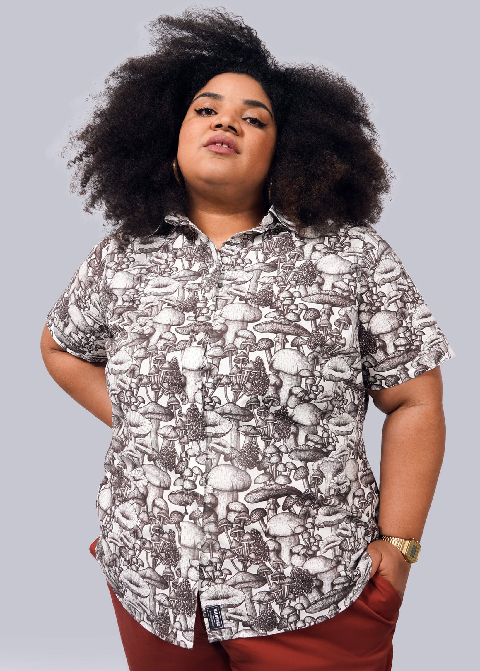 1X= A confident woman with curly hair wears The Essential Button Up, a short-sleeved, breathable cotton shirt with a black and white mushroom pattern. She pairs it with red pants and poses against a plain white background.