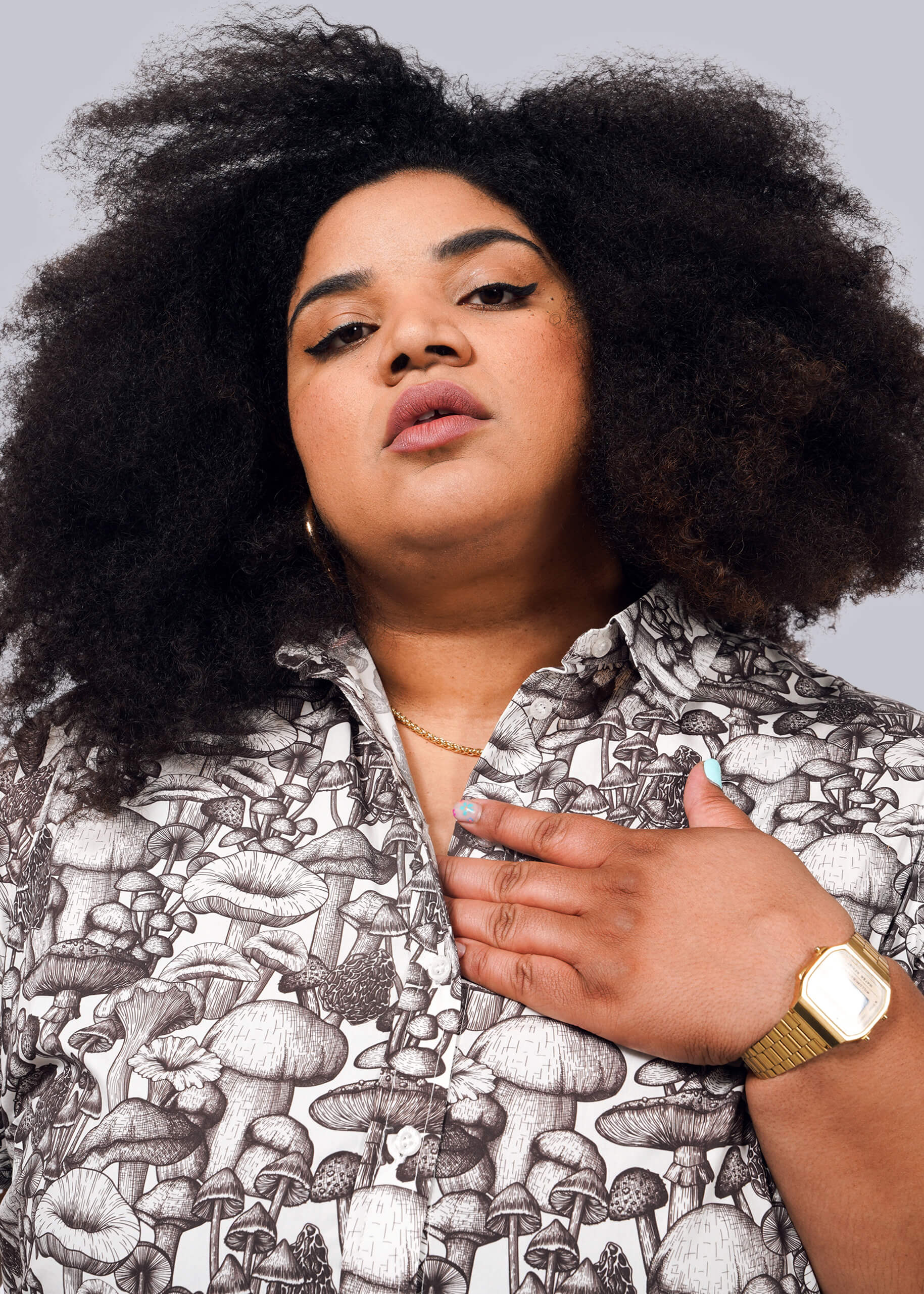 1X=A person with voluminous curly hair wears The Essential Button Up, featuring a mushroom pattern, styled with a gold watch, necklace, and earrings. They look confidently at the camera, one hand on their chest against a plain white background.