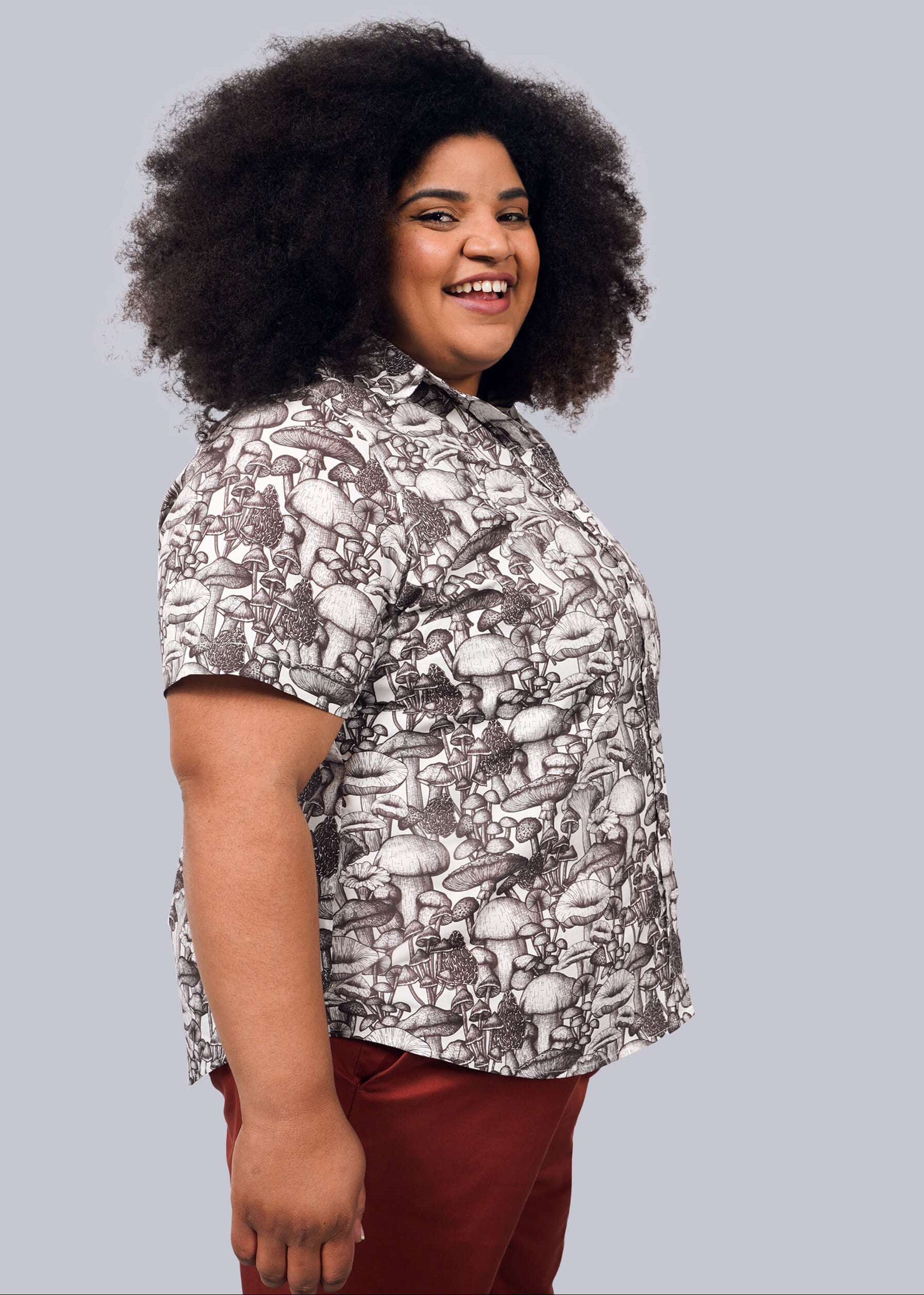1X= A person with voluminous curly hair smiles sideways, wearing The Essential Button Up—a short-sleeved, breathable cotton shirt with a mushroom pattern and Wildfang-branded buttons—paired with reddish-brown pants against a plain white background.