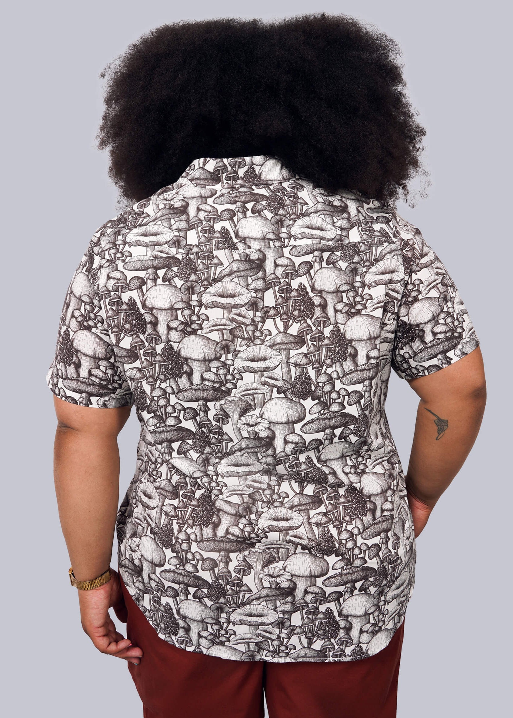 1X=Person with curly hair facing away, wearing The Essential Button Up— a short-sleeve shirt with a black and white mushroom pattern, Wildfang-branded buttons, and breathable cotton—paired with reddish-brown pants against a plain white background.
