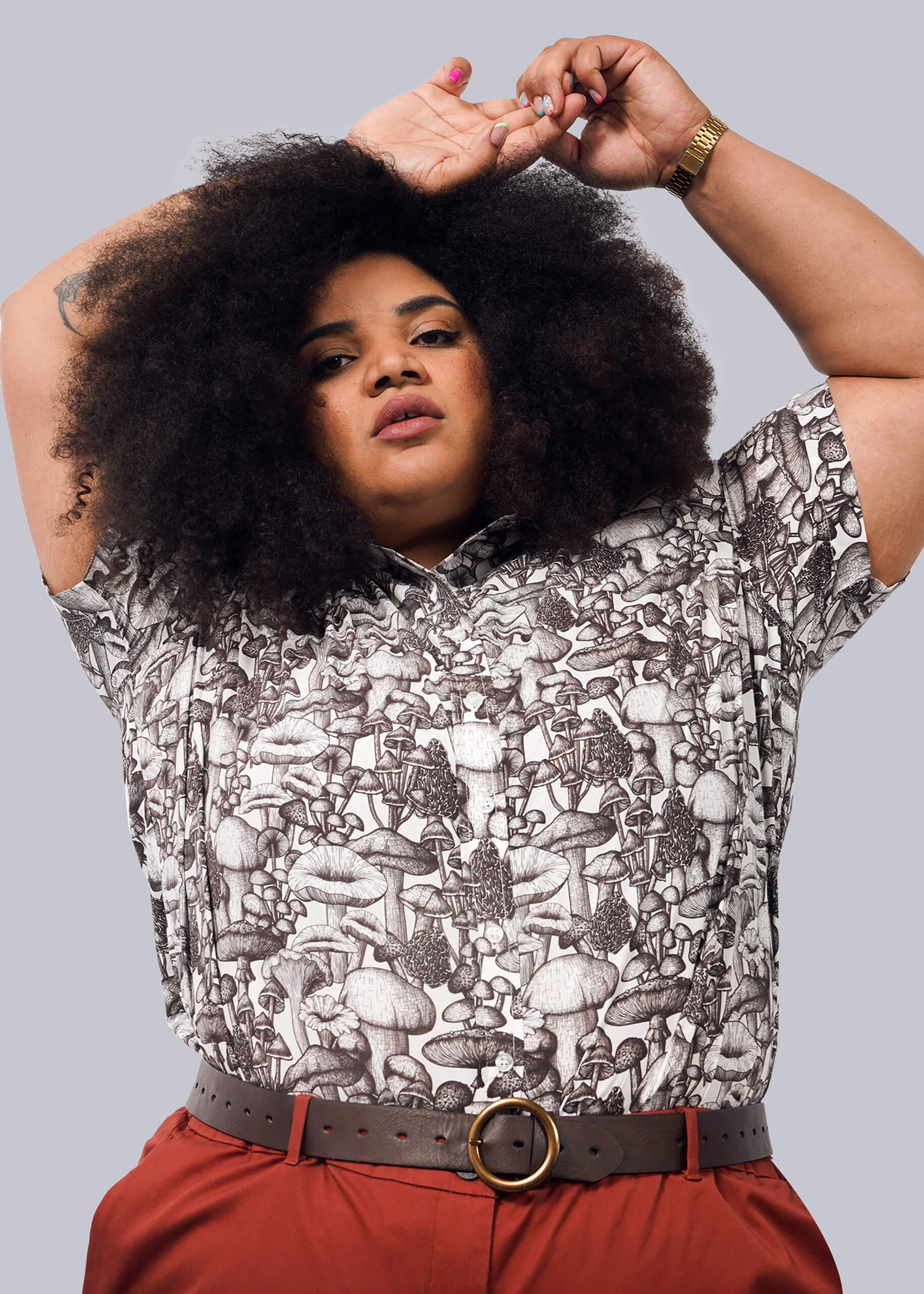 1X=A person with voluminous curly hair poses with arms raised, wearing The Essential Button Up—a patterned mushroom shirt made from breathable cotton with Wildfang branded buttons—along with red pants and a brown belt. Plain white background.