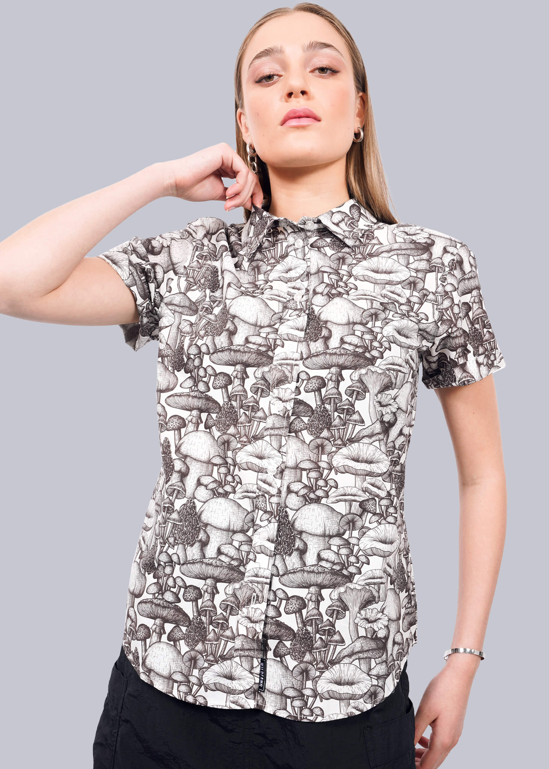 S=Someone stands against a white background wearing The Essential Button Up, a short-sleeve breathable cotton shirt with a detailed black-and-white mushroom print and Wildfang branded buttons, resting one hand on their neck and looking confidently at the camera.