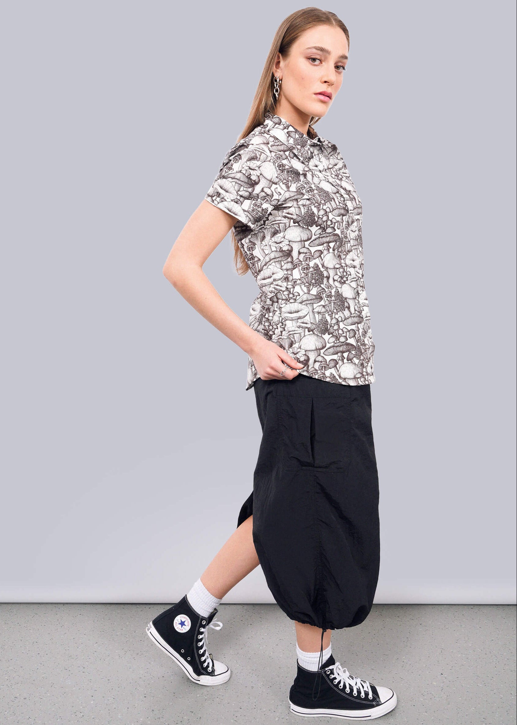 S=A young woman stands sideways against a white wall, wearing The Essential Button Up by Wildfang—a black and white patterned shirt with branded buttons—paired with a black midi skirt and black high-top Converse. She gazes confidently at the camera.