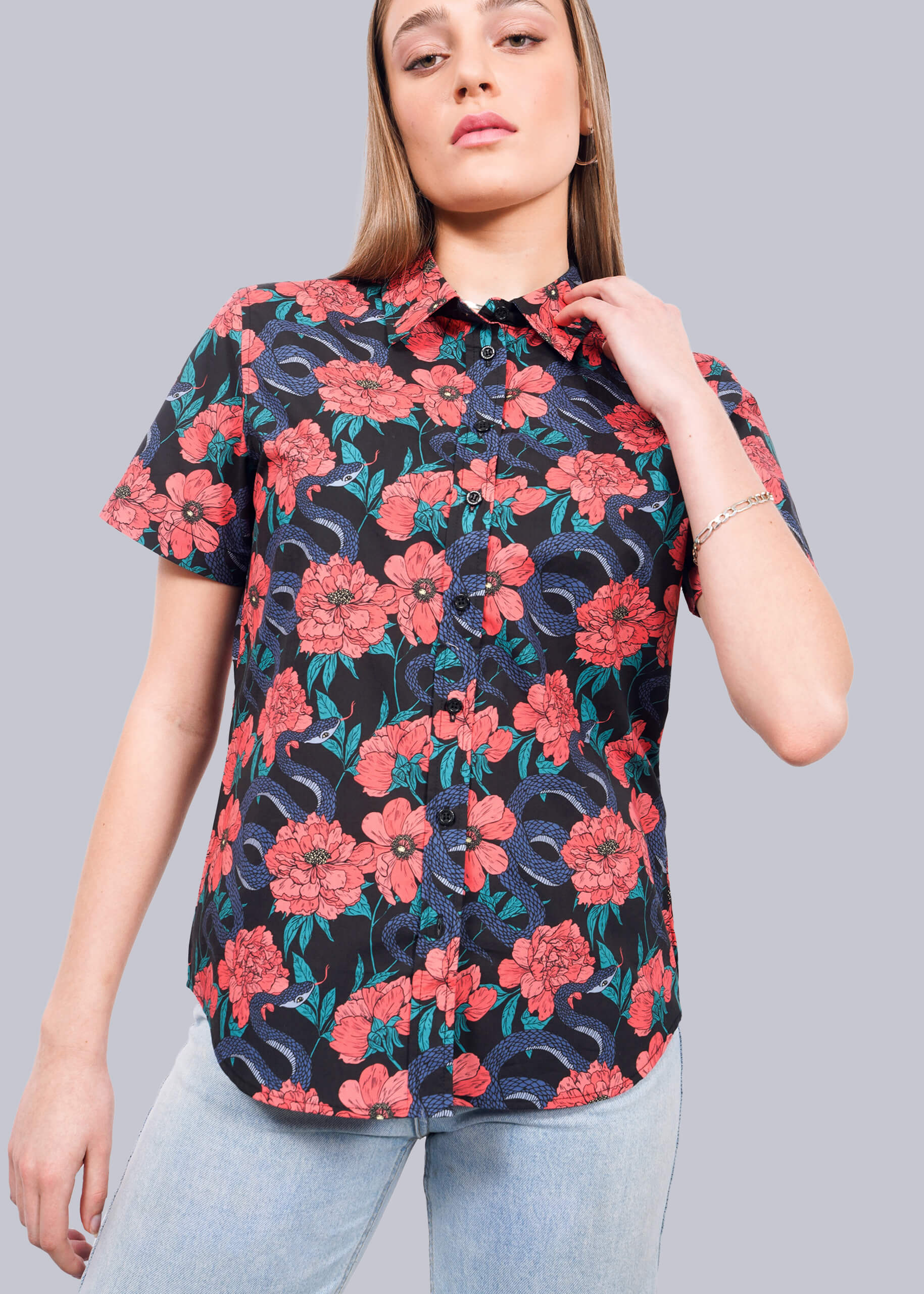 A woman wears The Essential Button Up by Wildfang, featuring a bold red, blue, and green floral pattern on breathable cotton, paired with light blue jeans as she poses against a plain white background.