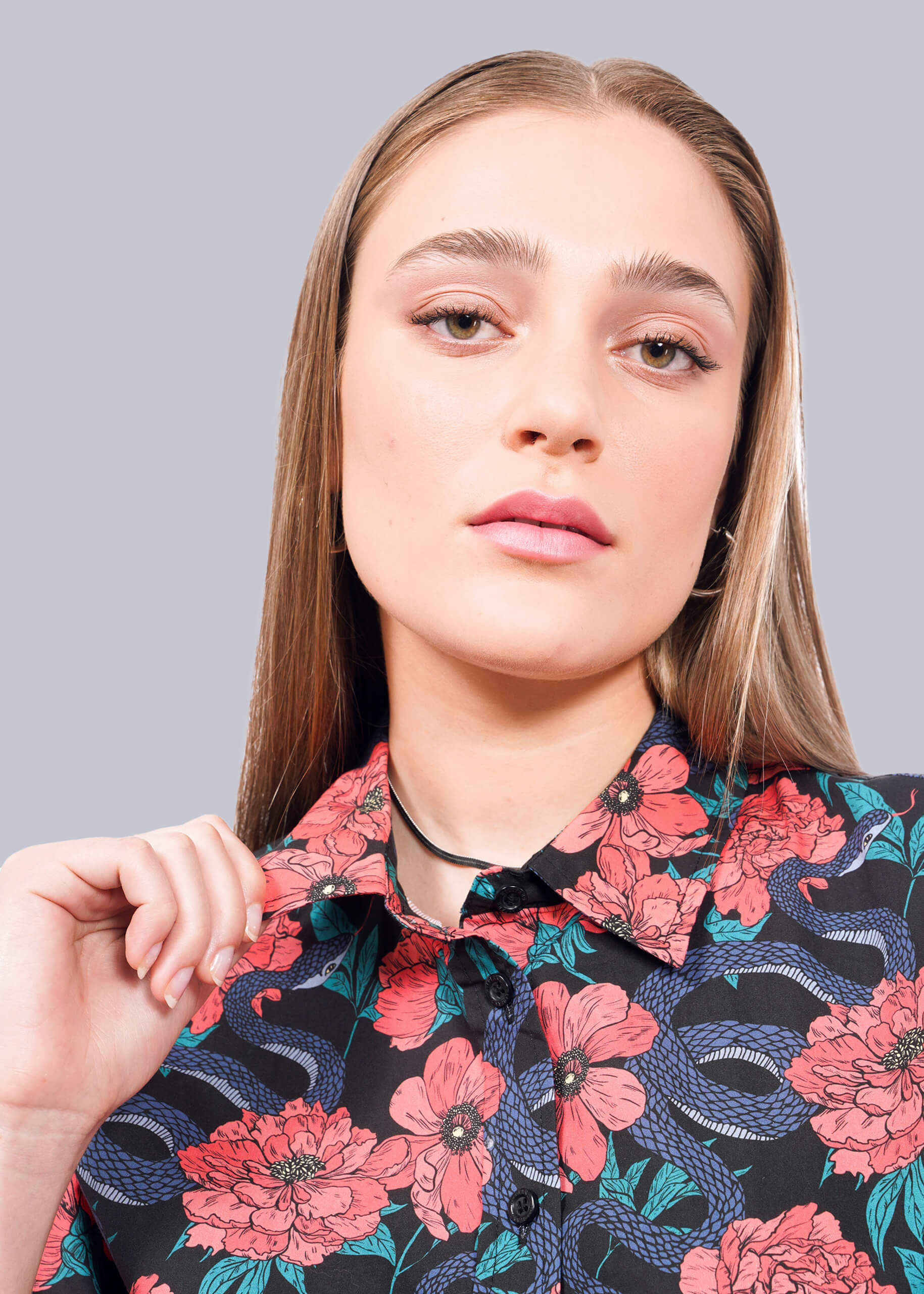 S=A young woman with straight, light brown hair wears The Essential Button Up, featuring pink floral and blue snake patterns. She holds the collar with one hand and looks confidently at the camera against a white background.