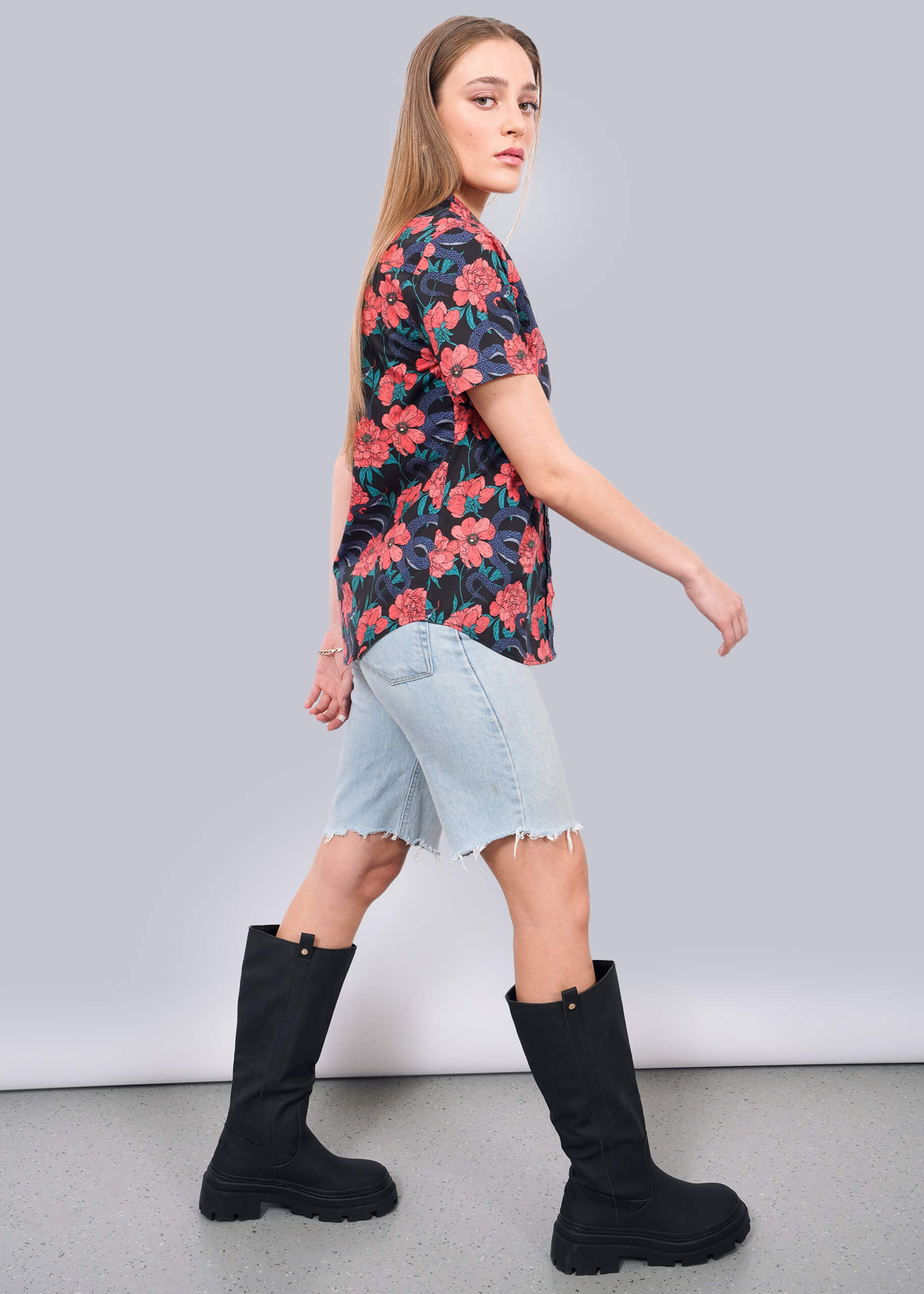 S=A woman with long blonde hair wears The Essential Button Up with a floral pattern, light denim knee-length shorts, and tall black boots, walking sideways against a plain white background.