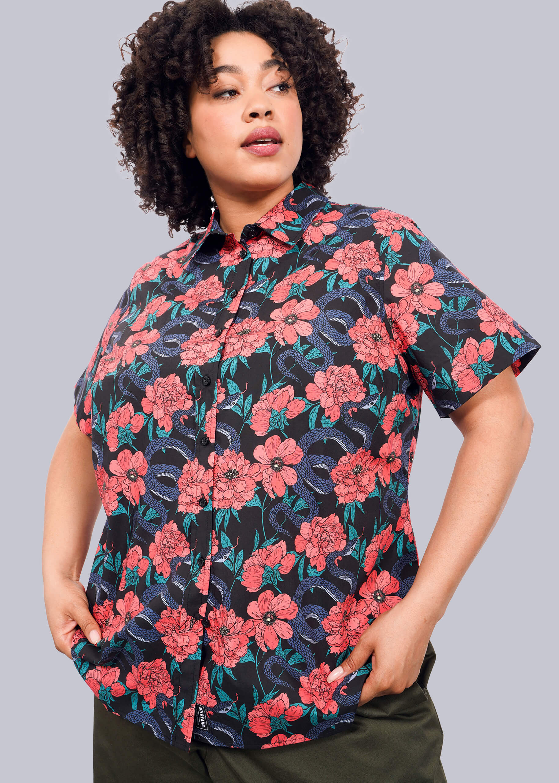 1X= A person with curly hair wears The Essential Button Up, a bright red and purple floral cotton shirt with a tailored fit, paired with dark green pants, posed against a plain white backdrop.