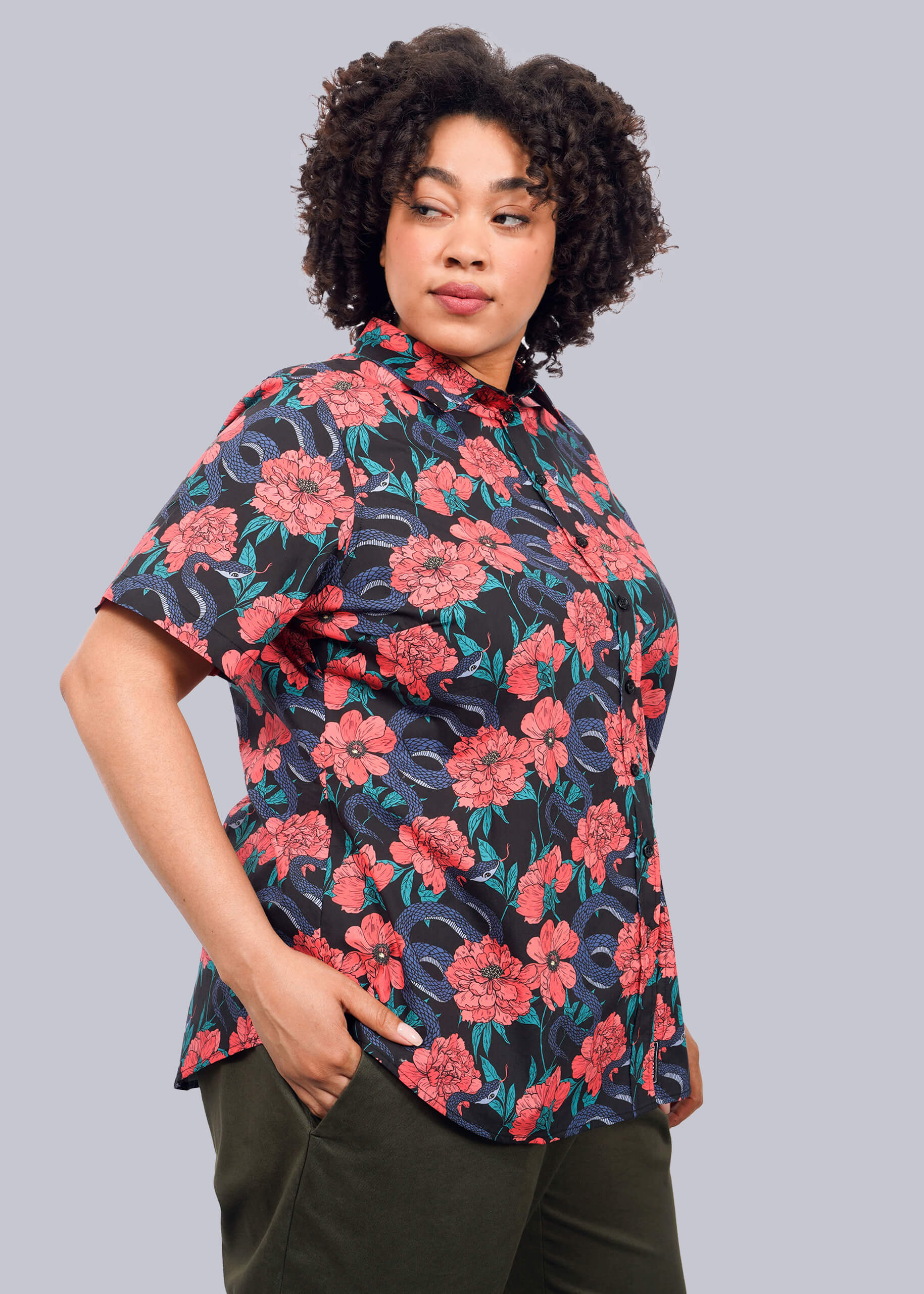 1X=A person with curly hair wears The Essential Button Up, tailored fit, short-sleeve, in a vibrant red and blue floral pattern, paired with dark green pants. They stand against a plain white background, looking to the side with hands in their pockets.