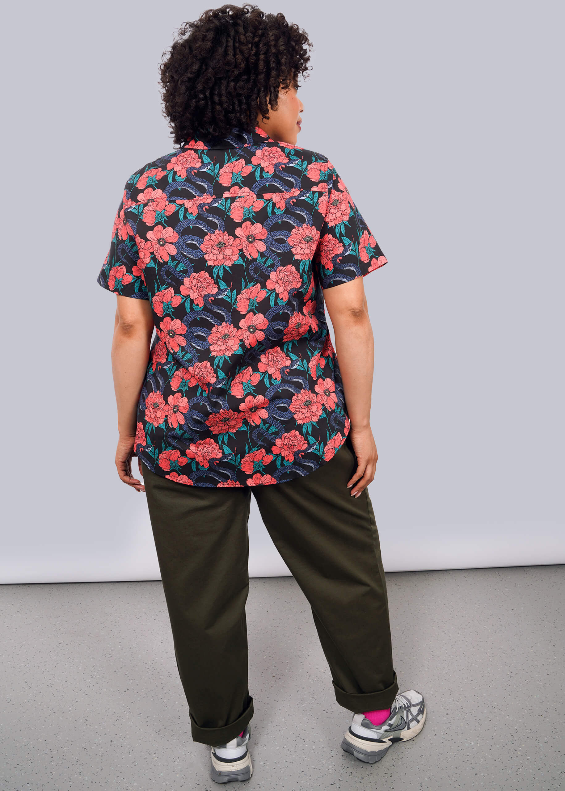 1X=A person with curly hair stands facing away, wearing The Essential Button Up—a bold red floral print short-sleeve shirt in breathable cotton—paired with olive green pants and sneakers. The background is plain white.