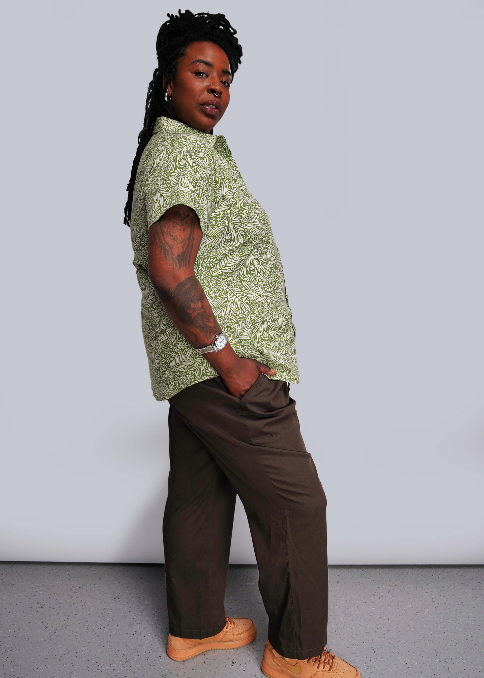 1X=Person wearing a green patterned shirt and brown pants against a plain background