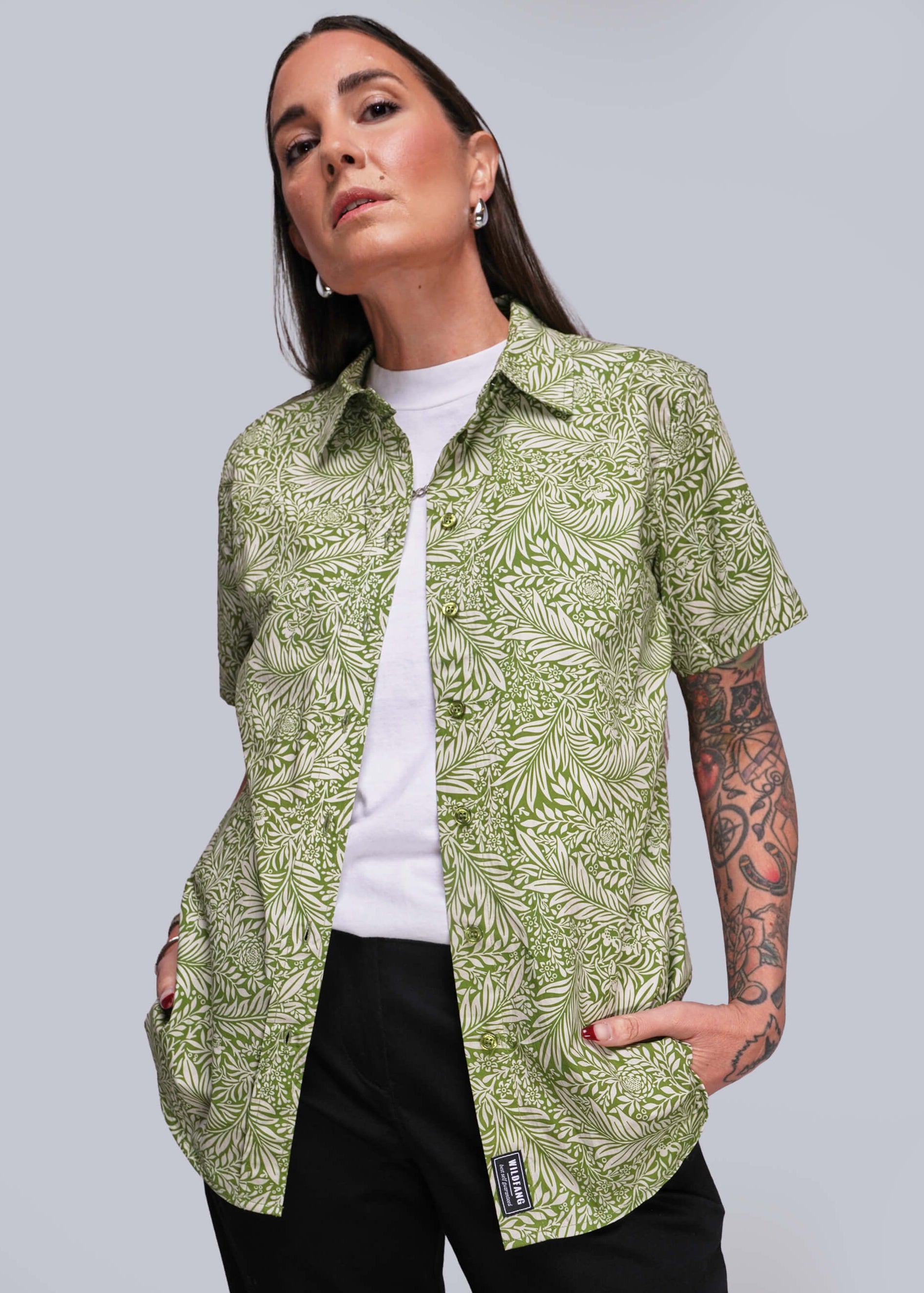 Person wearing a green patterned shirt over a white top with black pants on a plain background