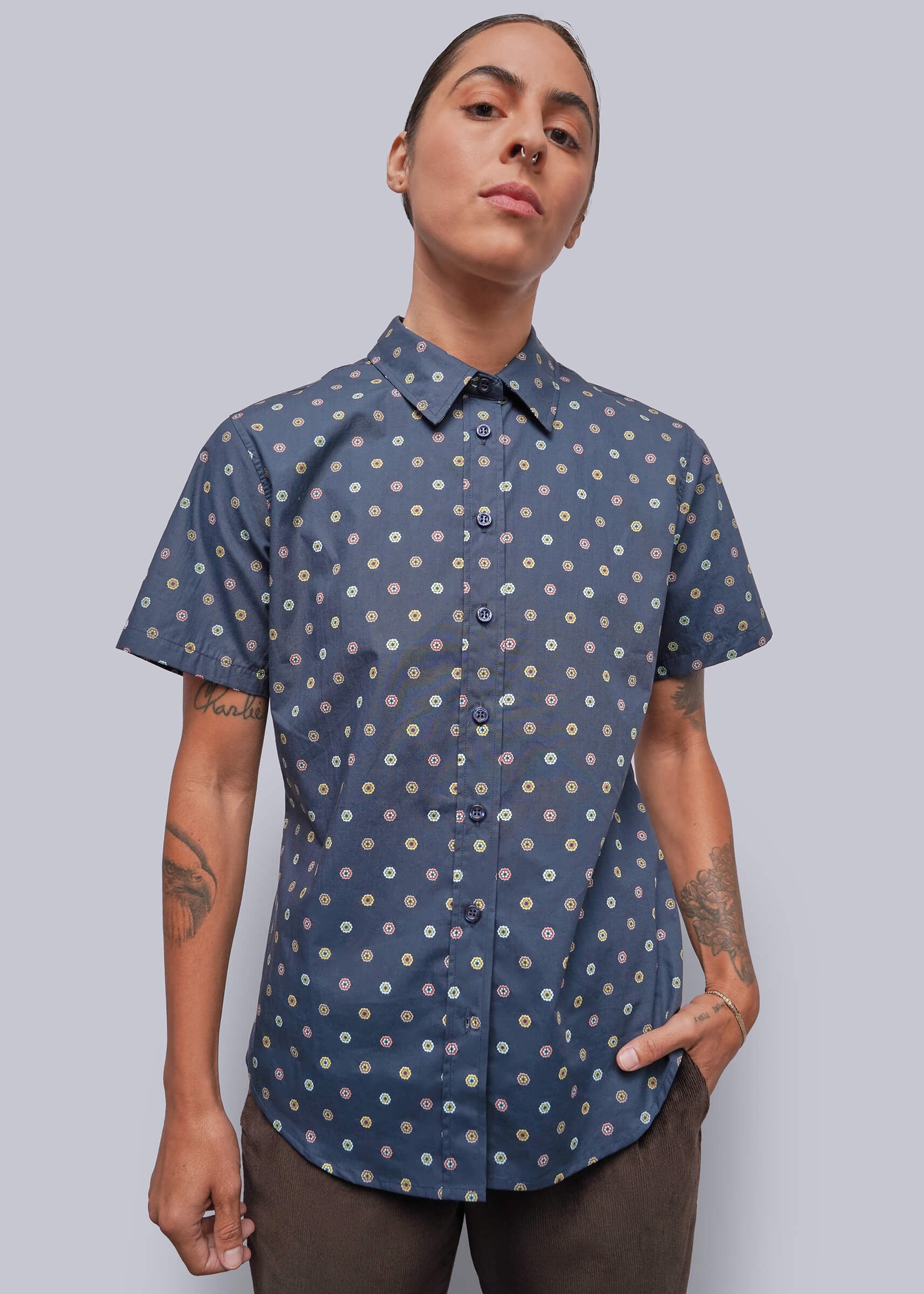 A model wearing a short-sleeve button-up shirt with a blue base and a small white and yellow floral pattern, paired with brown pants, against a solid gray background.