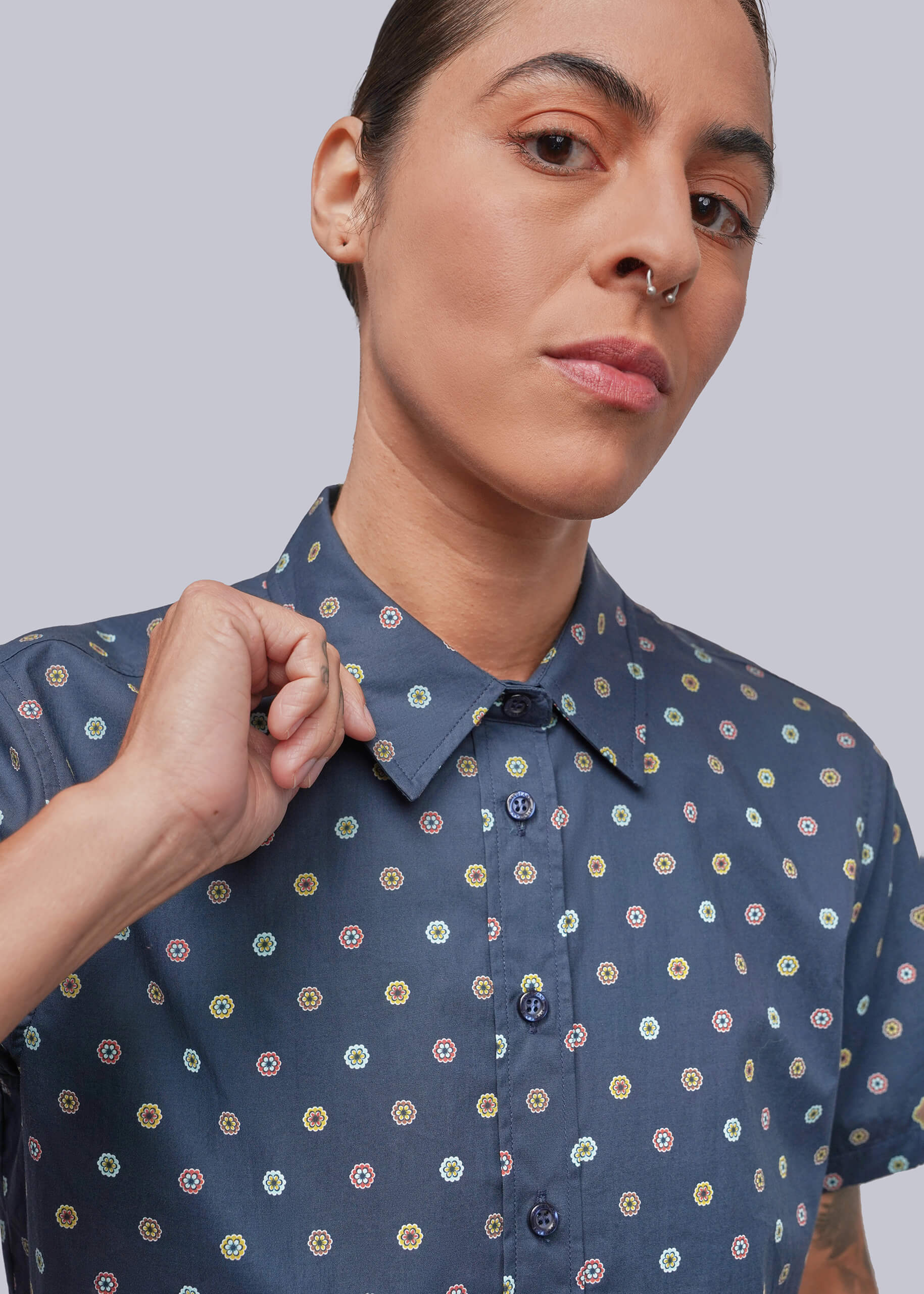 A close-up of a person wearing a short-sleeve button-up shirt in slate blue with a repeating multicolor circular pattern, set against a solid light gray background.