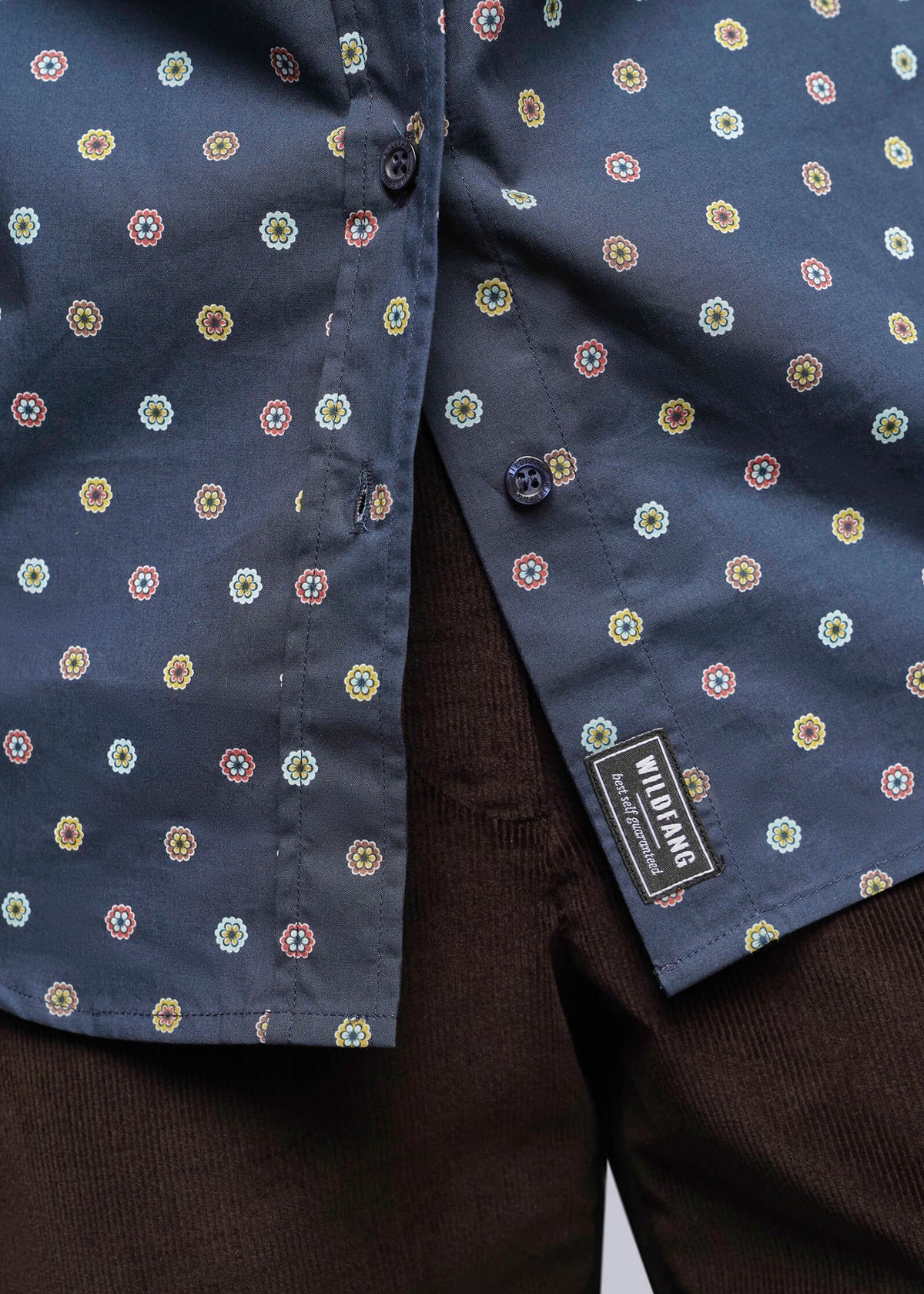 S=Close-up of a navy blue button-up shirt with a small multicolor geometric pattern, worn over dark brown corduroy trousers. A black brand label reading 'Mikrofashion' is visible near the hem.
