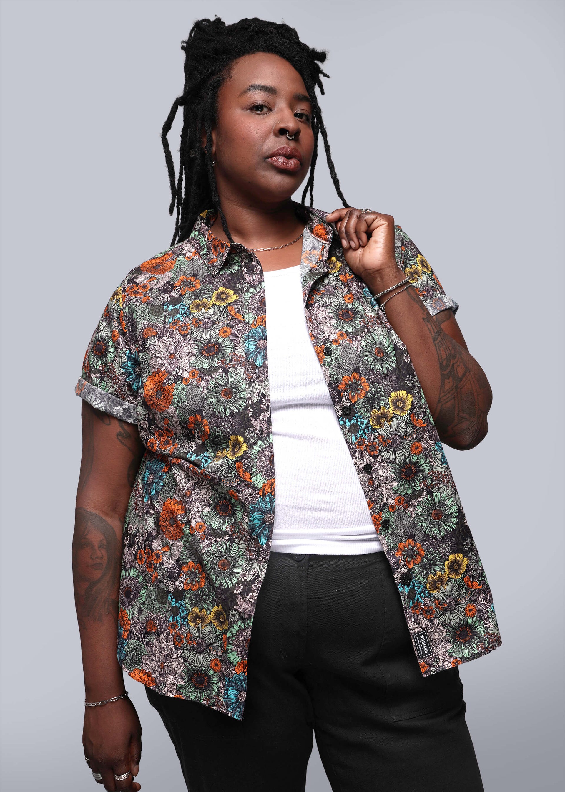 1X=Model wears white shirt under floral printed short sleeved button up shirt and black pants in front of grey background.