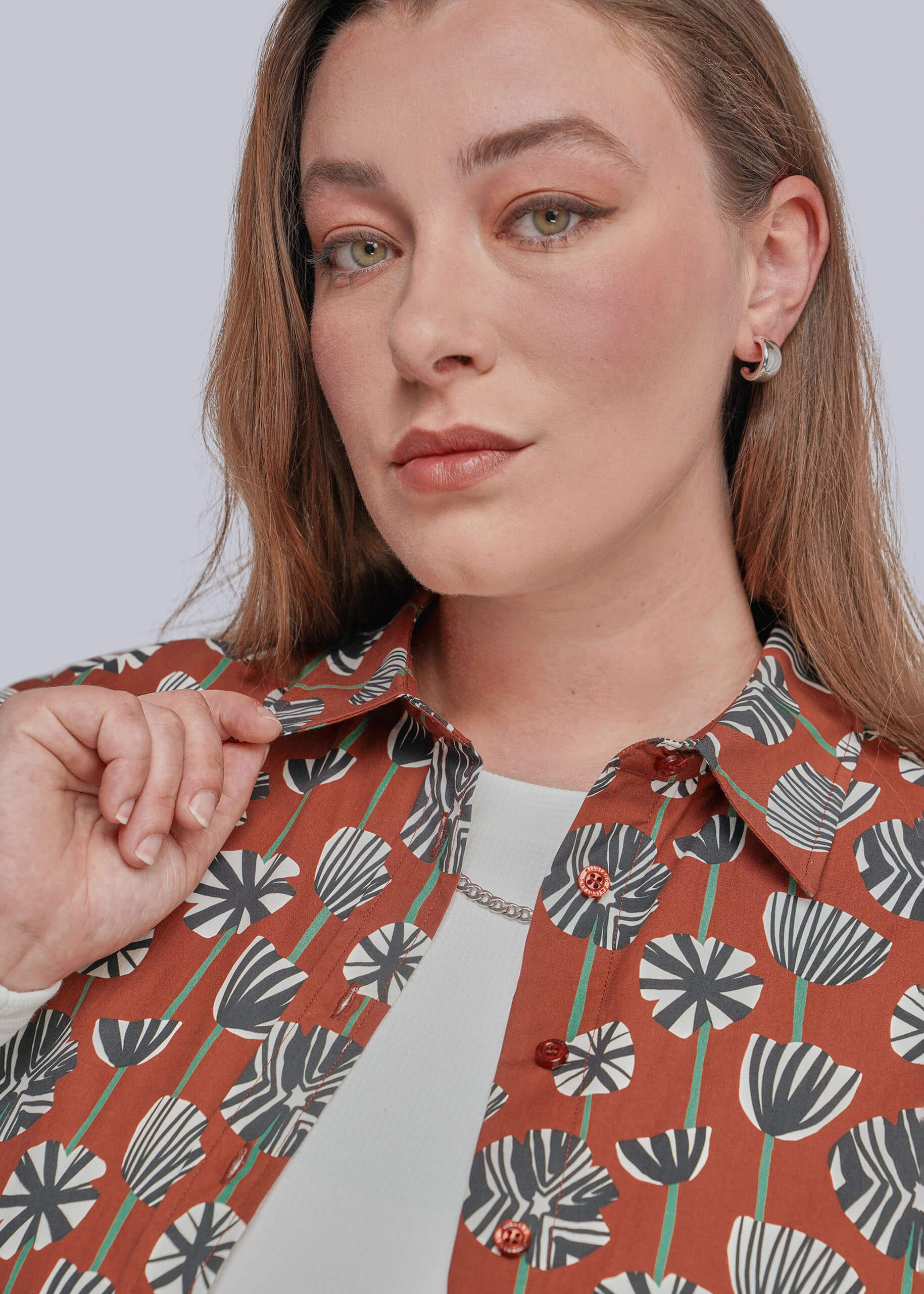 1X=A close-up of a woman wearing a rust-orange button-up shirt with a black, white, and green botanical leaf pattern, layered over a white top.
