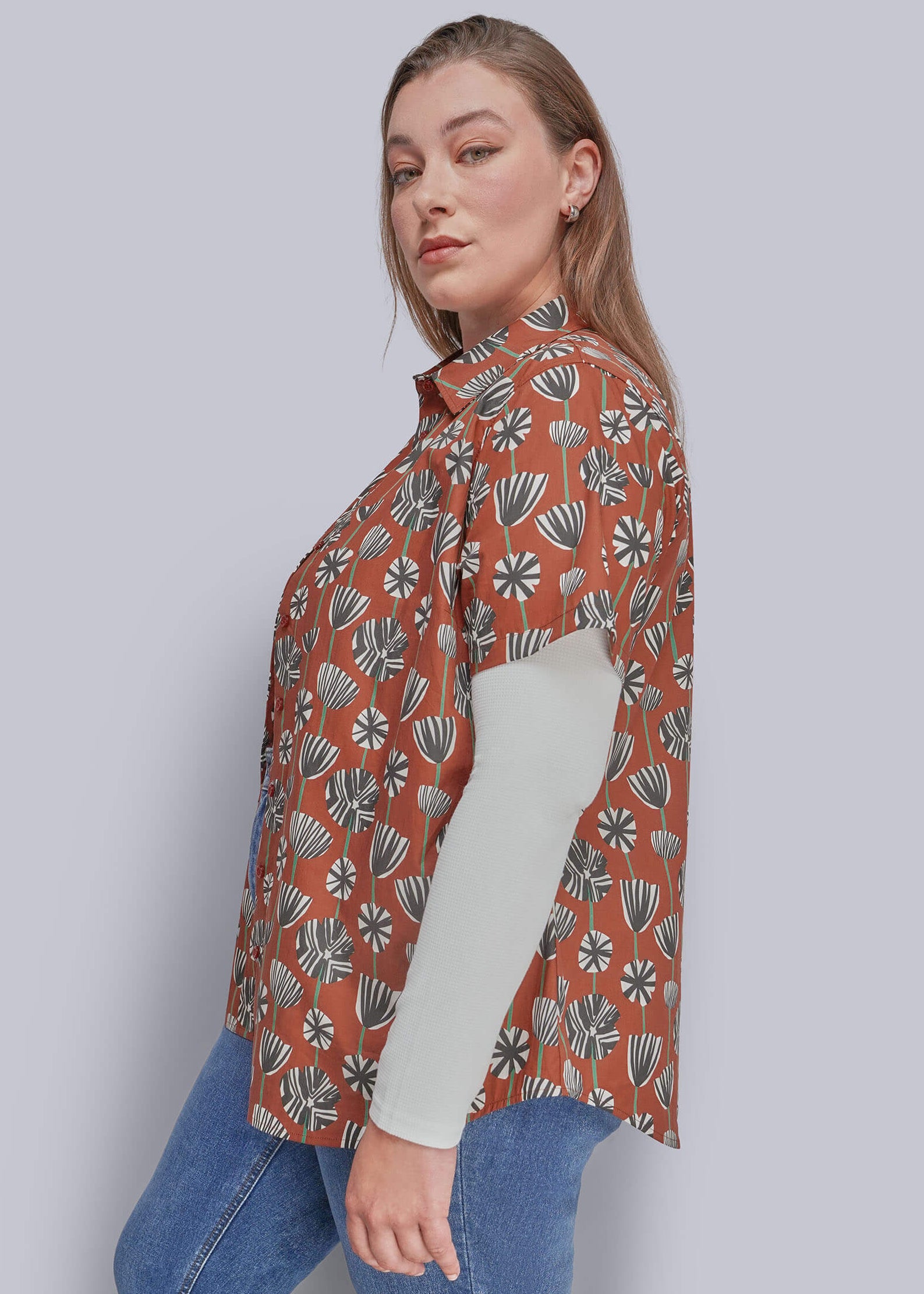 1X=A woman modeling a rust-orange short-sleeve button-up shirt with a white, gray, and green botanical pattern, layered over a light beige long-sleeve top and paired with blue jeans, against a solid gray studio background.