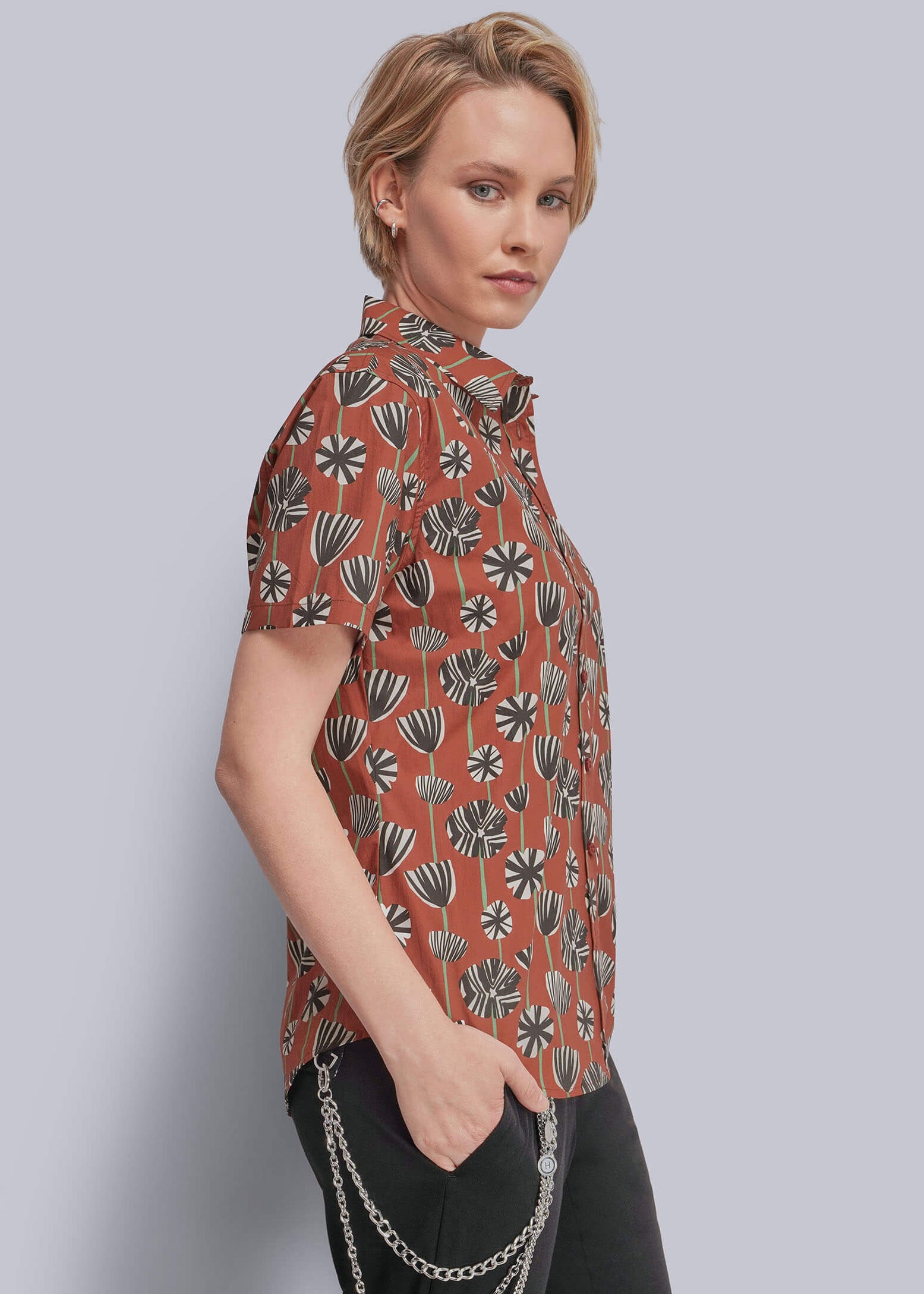 S=A woman modeling a short-sleeve button-up shirt with a rust-orange base and a black, gray, and white geometric floral pattern, paired with black pants and a silver chain accessory.