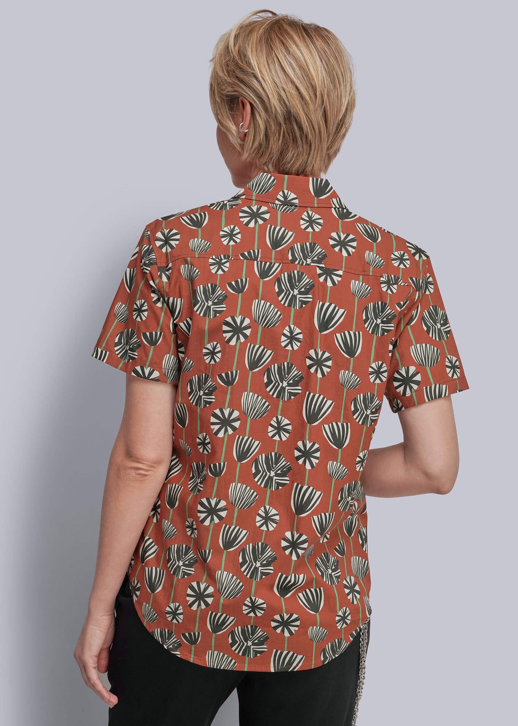 S=A woman shown from the back wearing a short-sleeve button-up shirt with a rust-orange base and a black and white botanical leaf pattern, paired with black pants, against a solid gray background.