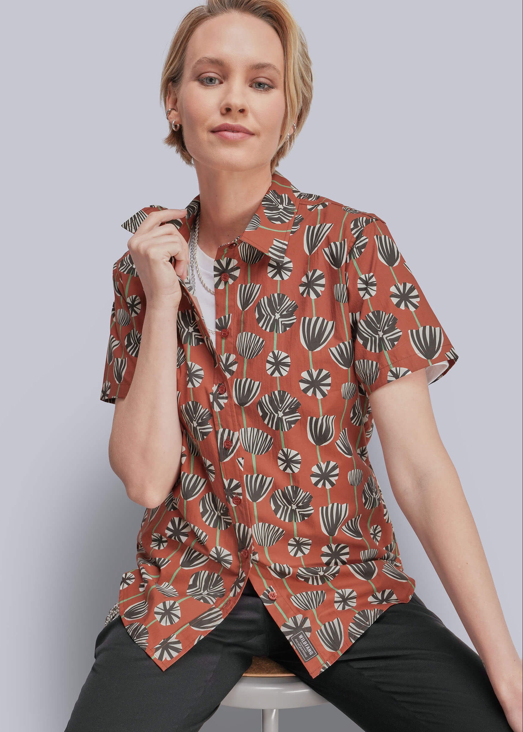 S=A woman wearing a short-sleeve button-up shirt with a terracotta orange base and a black and gray botanical pattern, paired with black pants, seated on a stool against a solid gray background.