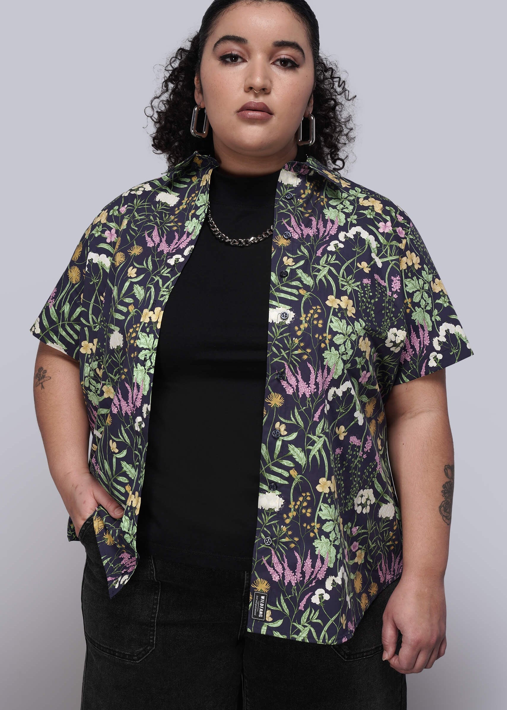 1X=Person wearing a colorful floral button up unbuttoned over a black top on a plain background
