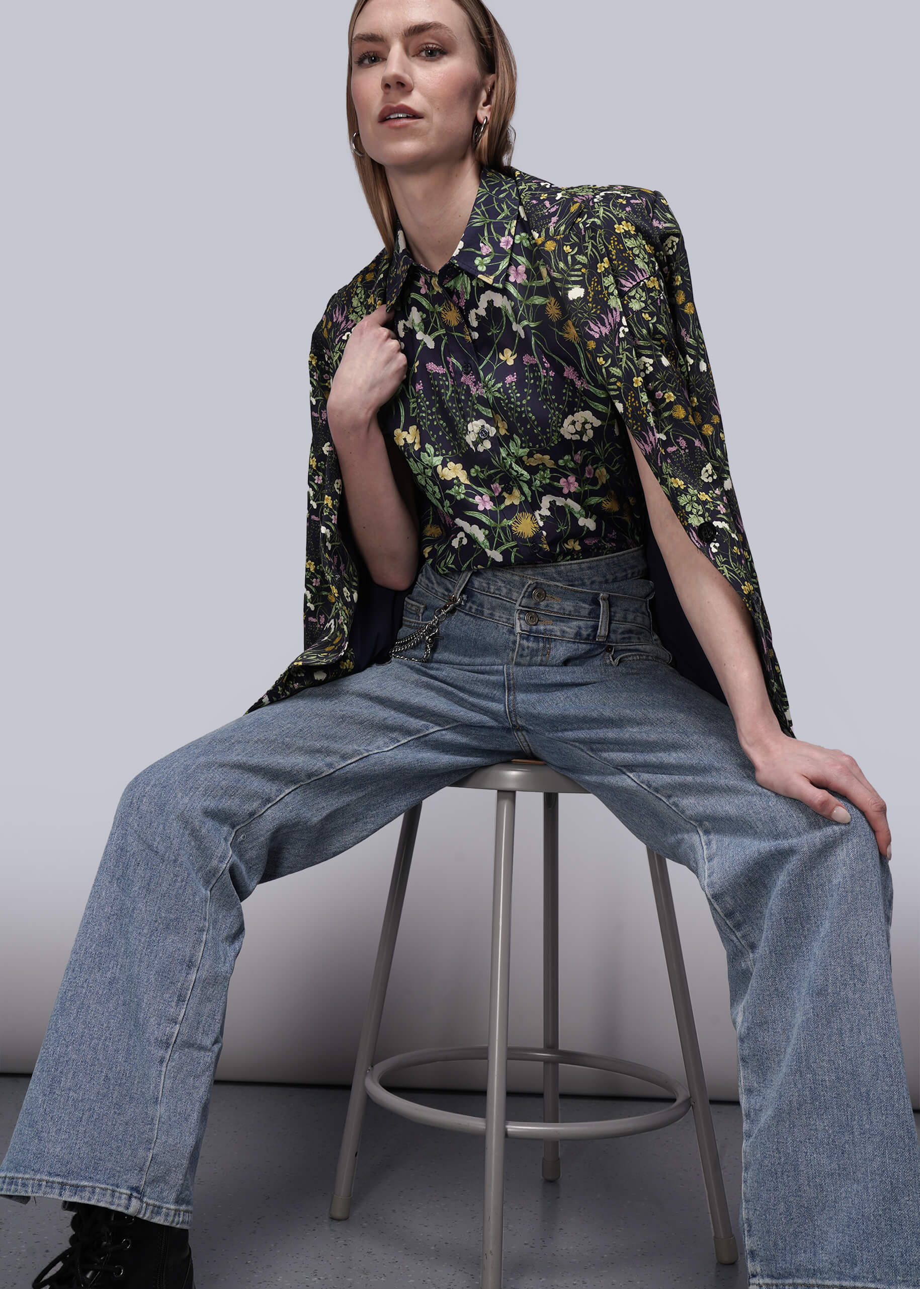 S=Person wearing a floral shirt, blazer, and jeans sitting on a stool against a plain background