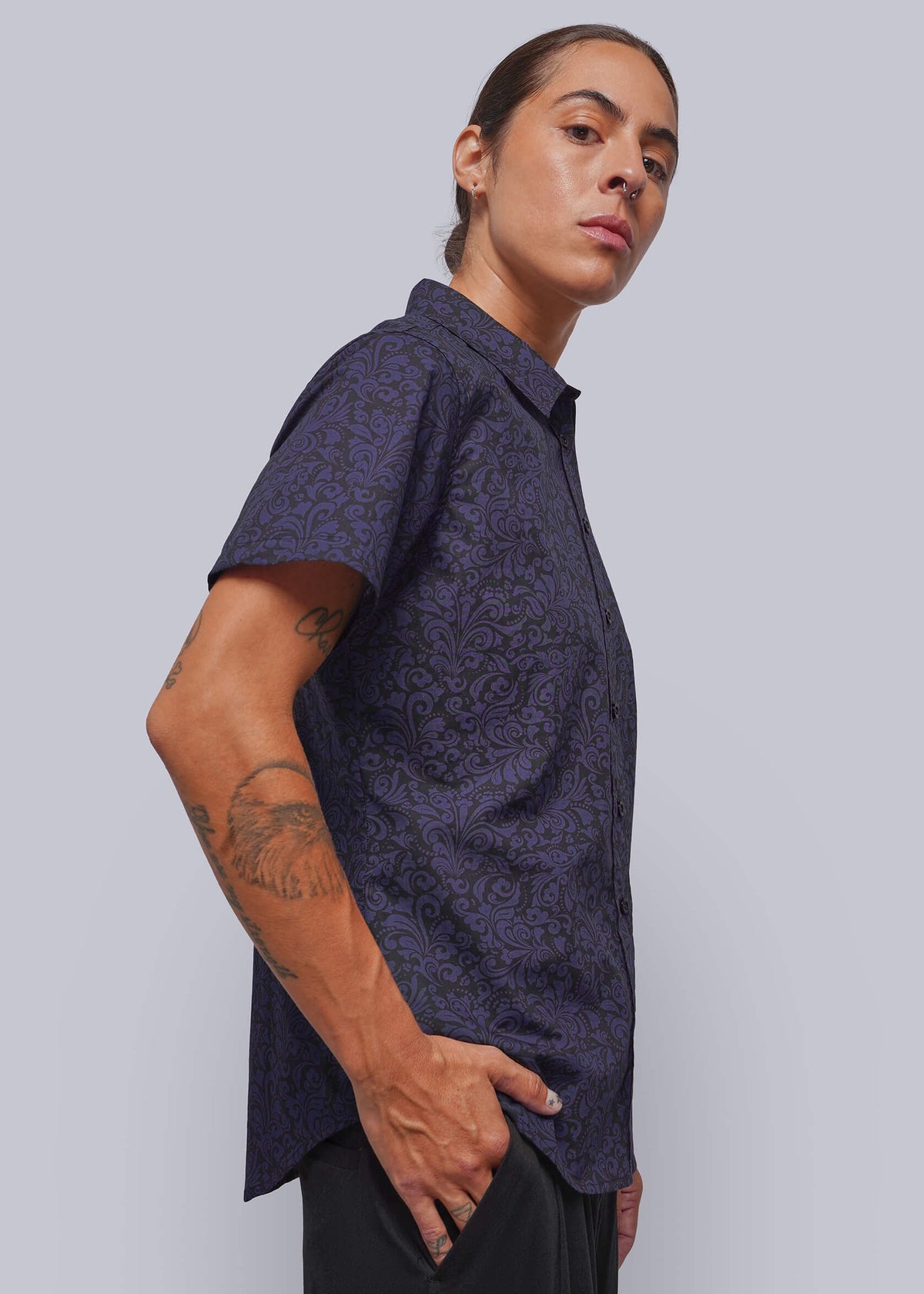 S=A model wearing a dark purple short-sleeve button-up shirt with a subtle paisley pattern, posed against a solid light gray background.