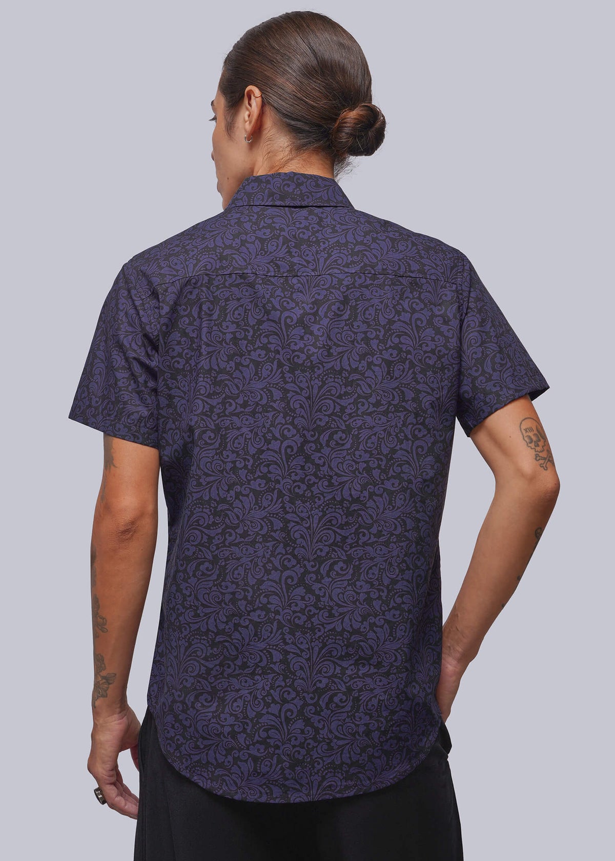 S=A person viewed from the back wearing a short-sleeve button-up shirt with an all-over dark purple and black damask pattern, paired with black trousers, against a solid light gray background.
