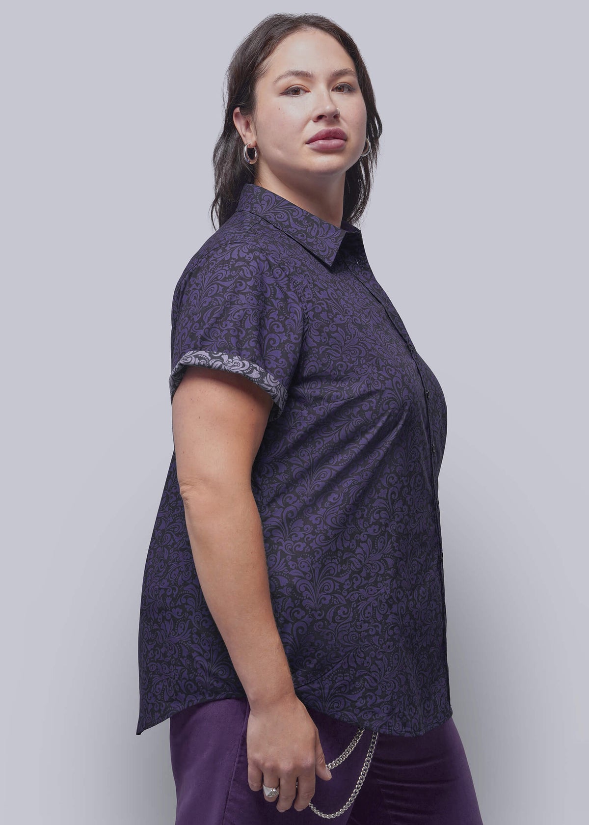 1X=A woman modeling a short-sleeve button-up shirt in dark purple with a subtle tonal floral pattern and a contrasting patterned cuff, paired with matching purple pants against a light gray studio background.