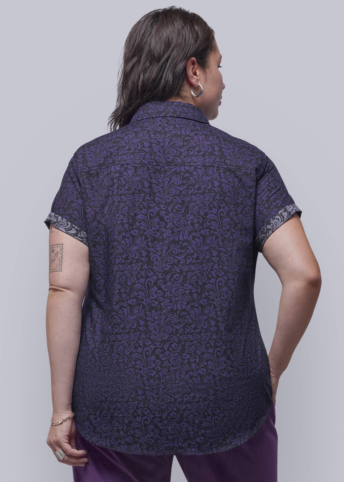1X=A person shown from the back wearing a short-sleeve button-up shirt with an all-over dark purple pattern and matching purple pants, photographed against a plain light gray studio background.